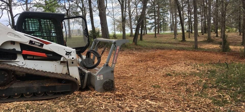 A white Bobcat skid-steer loader with a mulching head parked on a bed of wood chips in a wooded area.