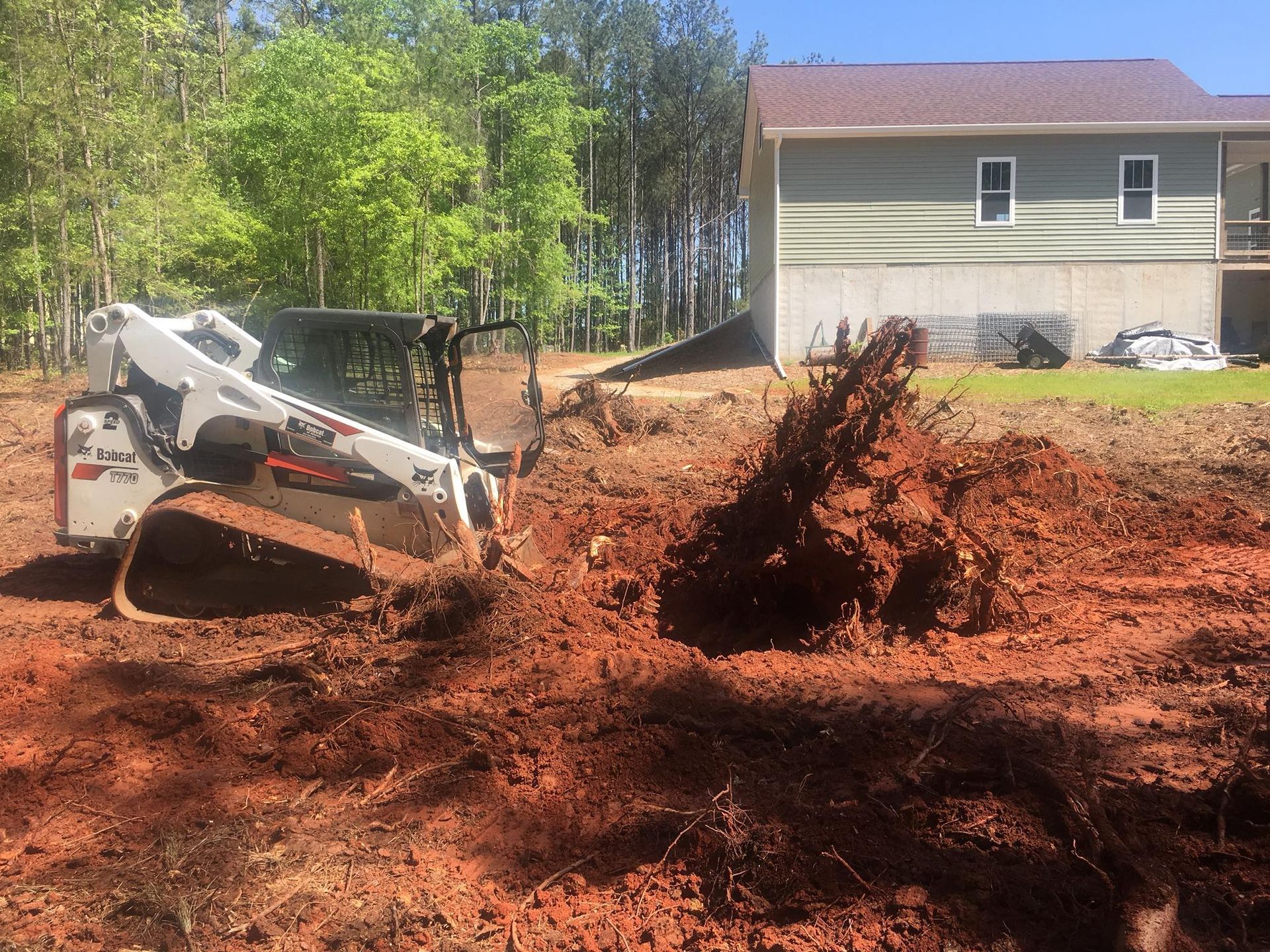 A white Bobcat skid-steer loader clears a large tree root and red dirt from a construction site near a house.