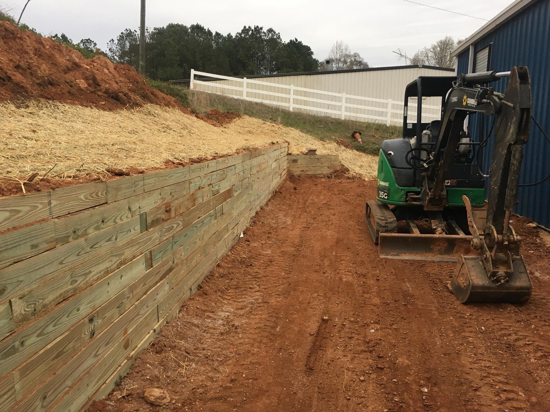 A green and black compact excavator sits near a newly constructed wooden retaining wall beside a building.