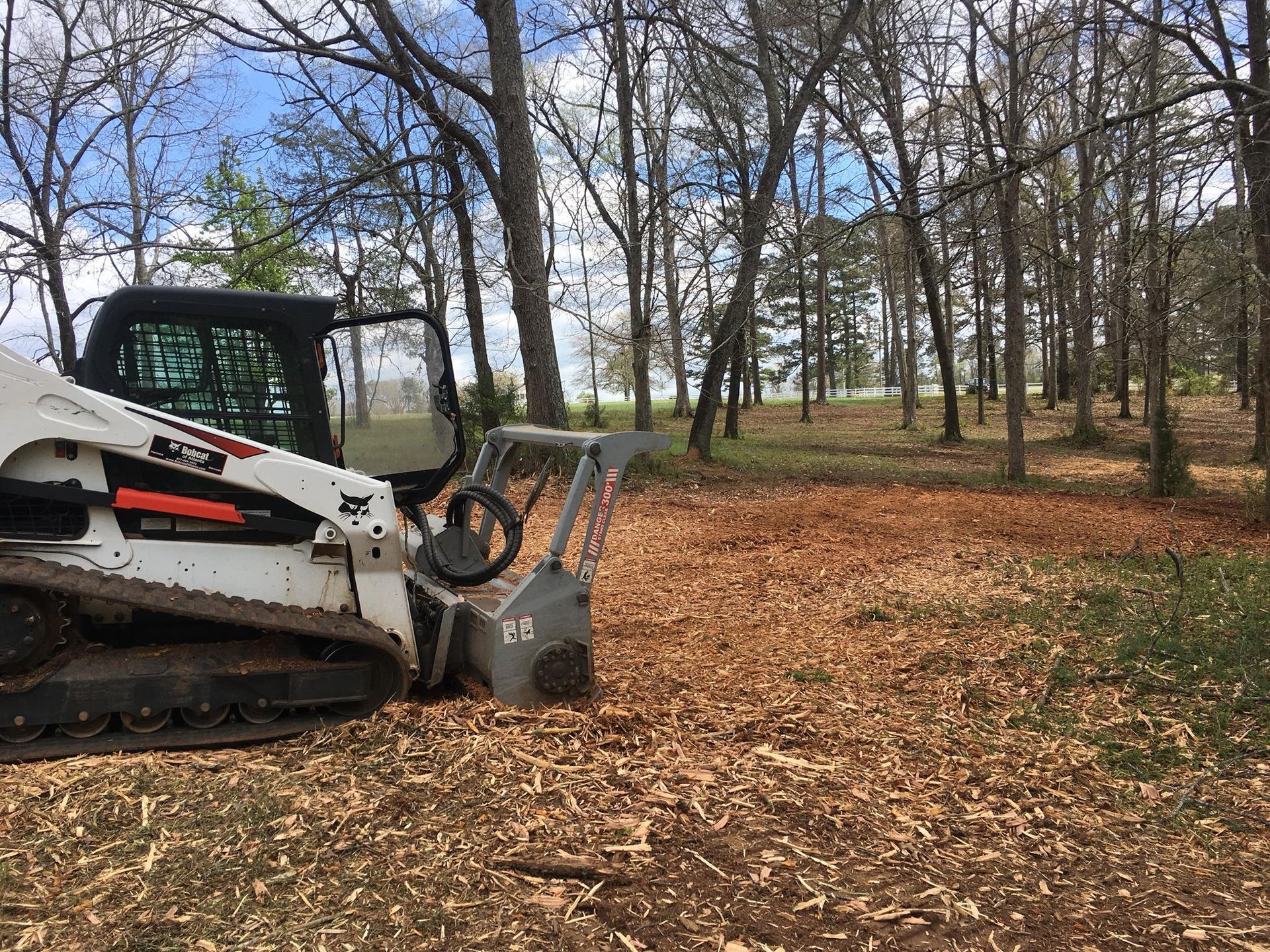 A white Bobcat skid-steer loader with a forestry mulcher attachment parked in a wooded area covered in wood chips.