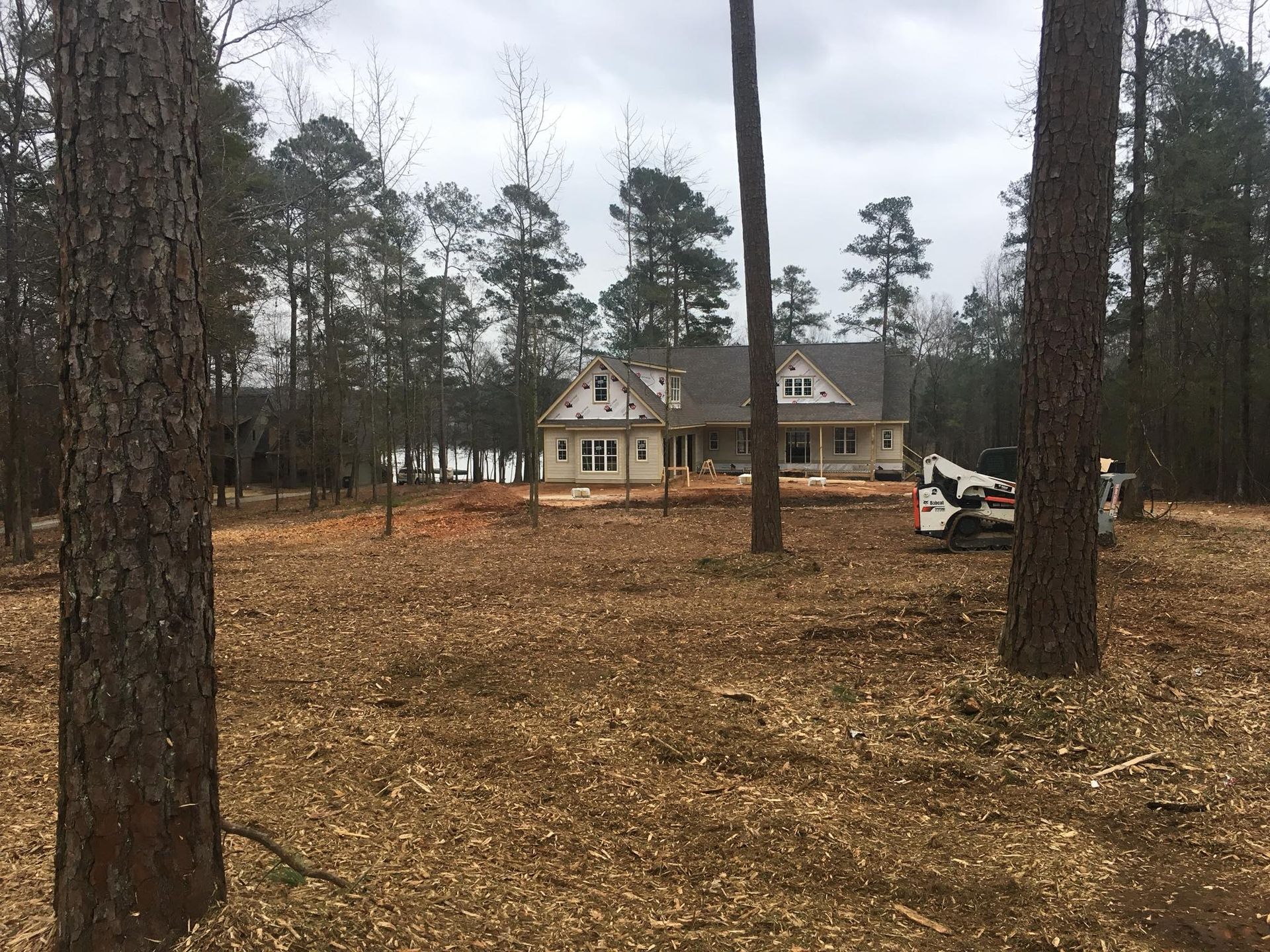 A newly built house under construction in a wooded lot with scattered leaves and a piece of heavy equipment nearby.