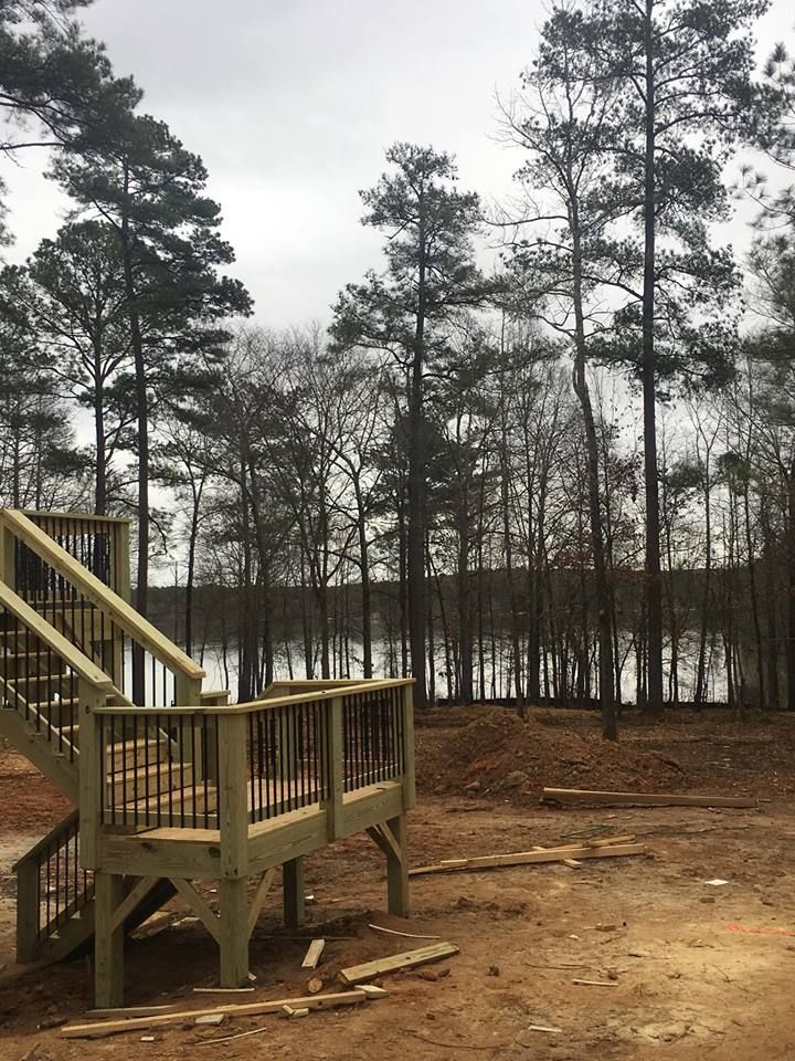 A newly constructed wooden deck and staircase stand on a dirt lot overlooking a lake surrounded by trees.
