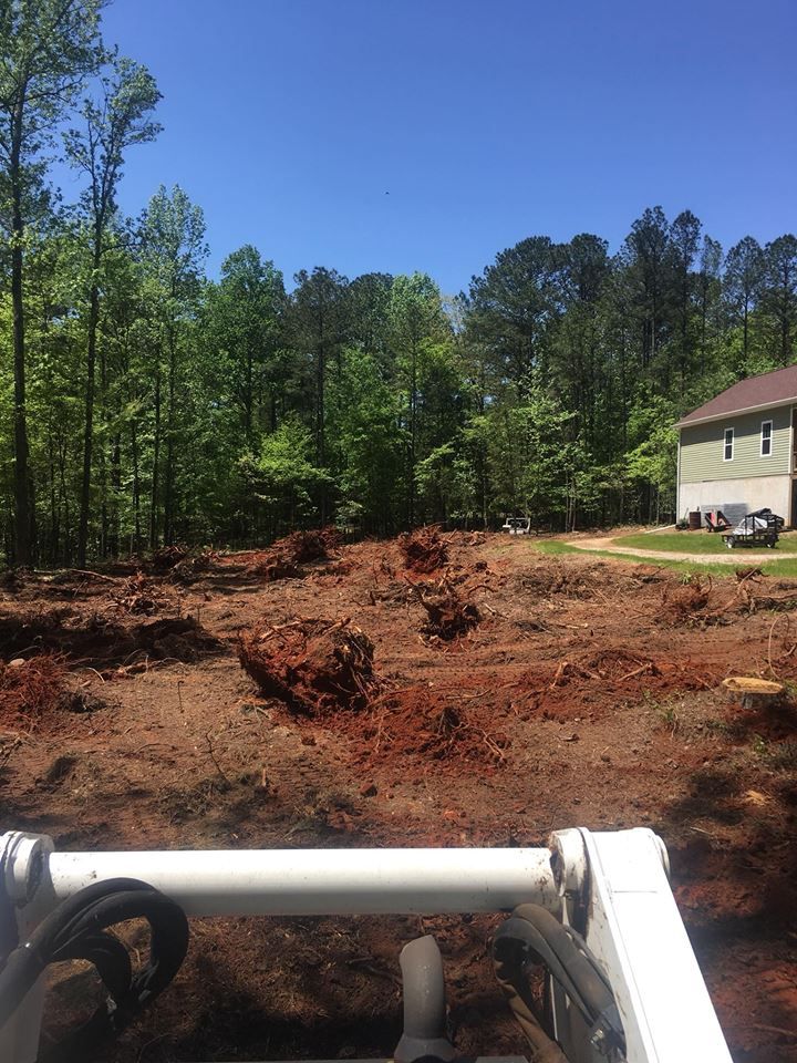 View from the cab of heavy machinery overlooking a cleared, uneven red dirt construction site near a wooded area and house.