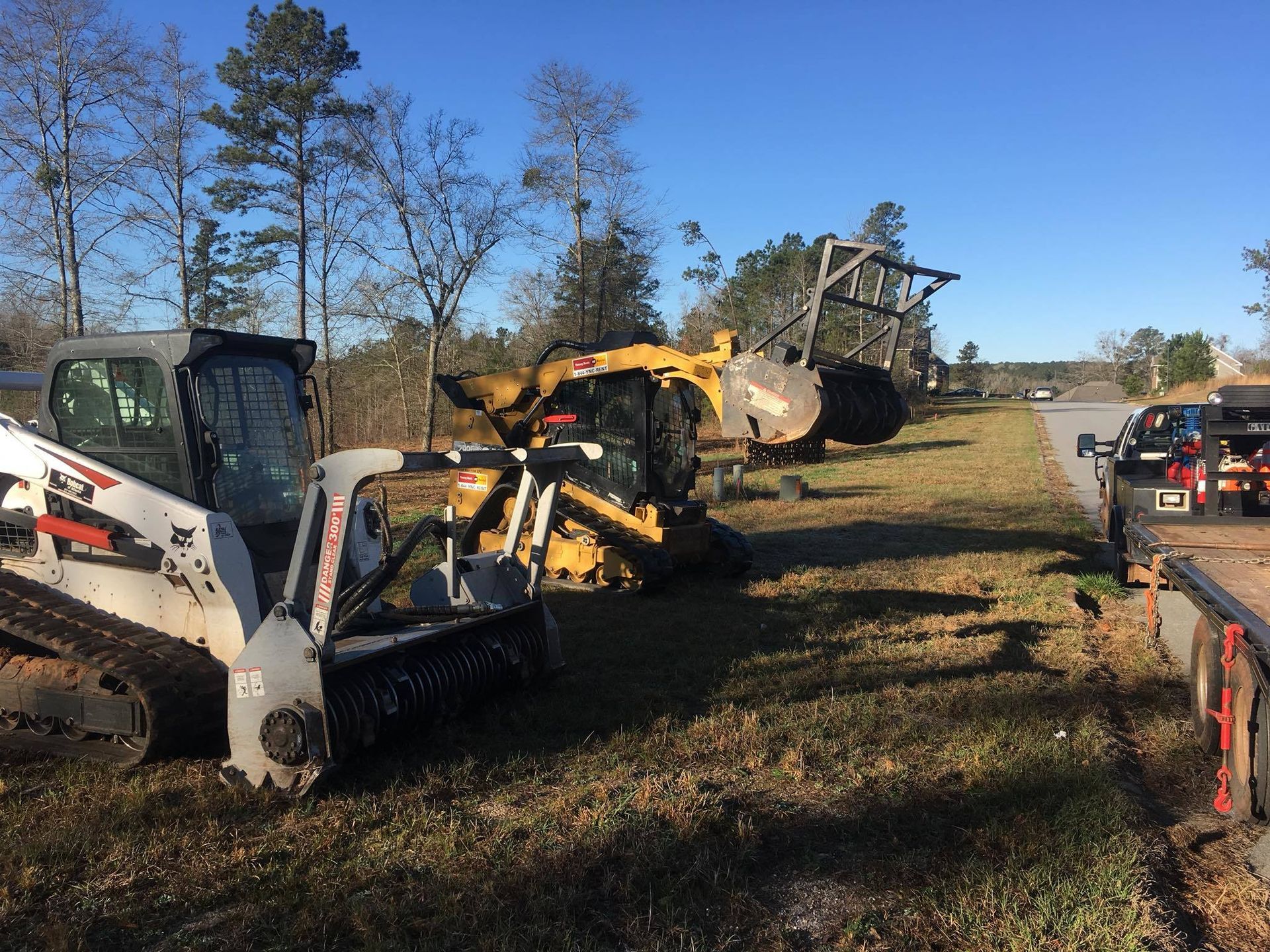 A white and a yellow skid steer loader with forestry mulcher attachments parked in a grassy field near a road.
