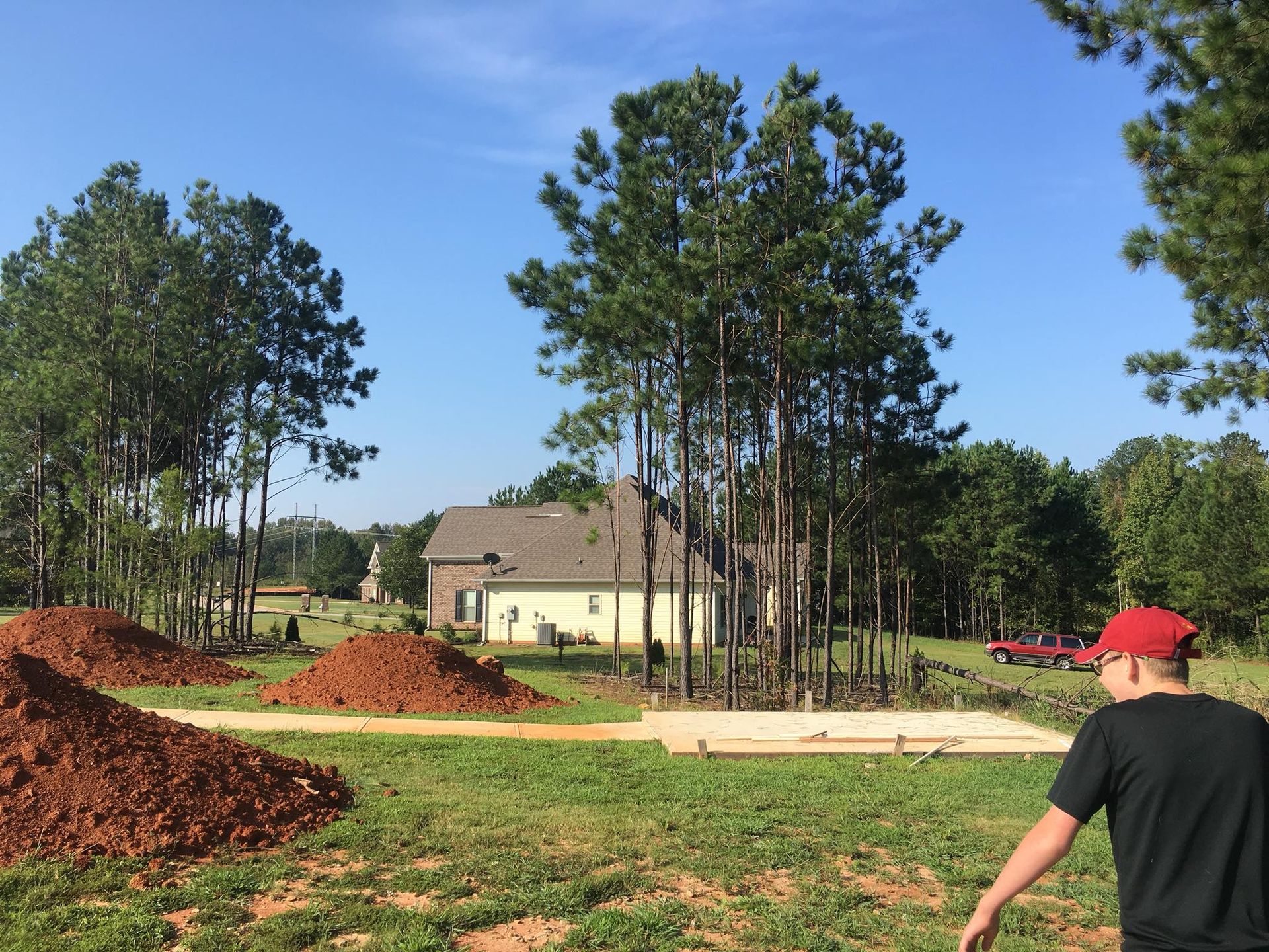 A person stands in a grassy lot with mounds of red dirt and a rectangular construction foundation near tall pine trees.