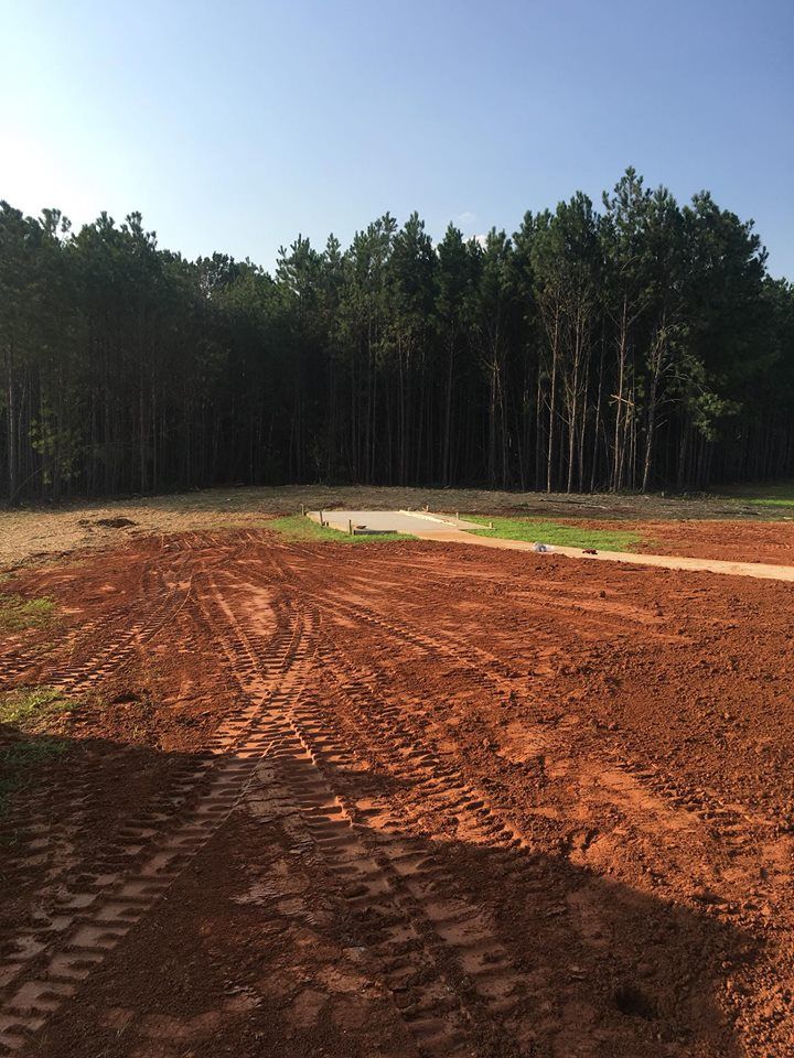 A construction site with reddish-brown soil, deep tire tracks, and a cleared path leading to a dense forest.