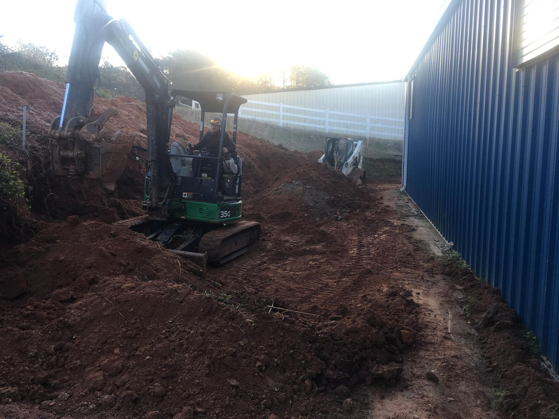An excavator digs in a dirt lot beside a blue, corrugated metal building at sunset.
