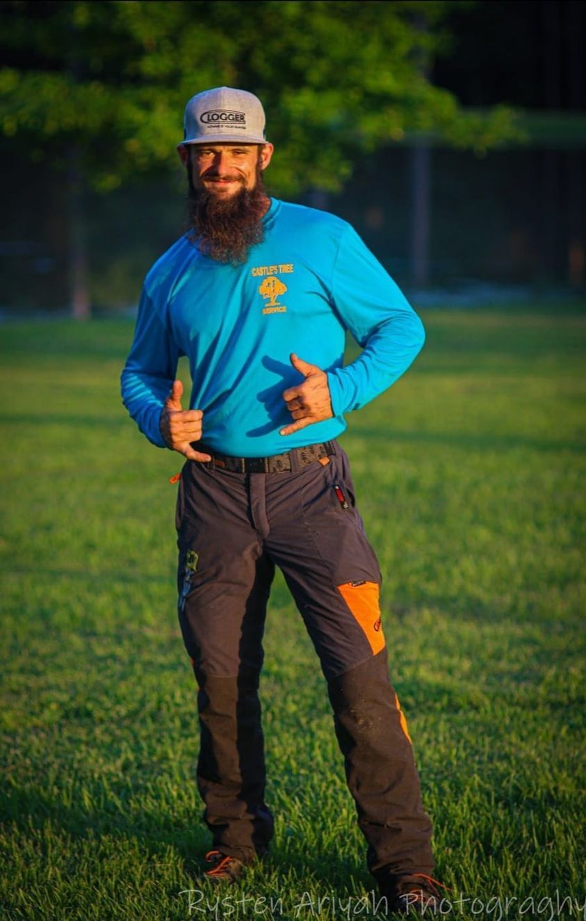 A man with a beard wearing a blue shirt and a hat is standing in the grass giving a thumbs up.