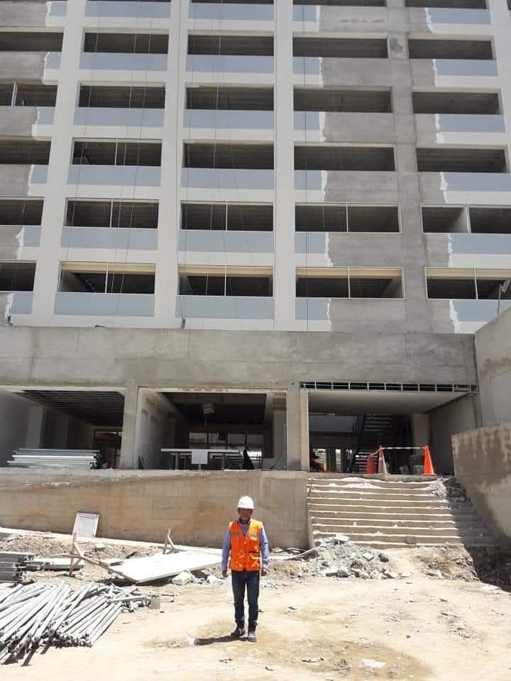 Un obrero camina hacia un edificio en construcción. Una estructura de hormigón gris con hileras de ventanas.