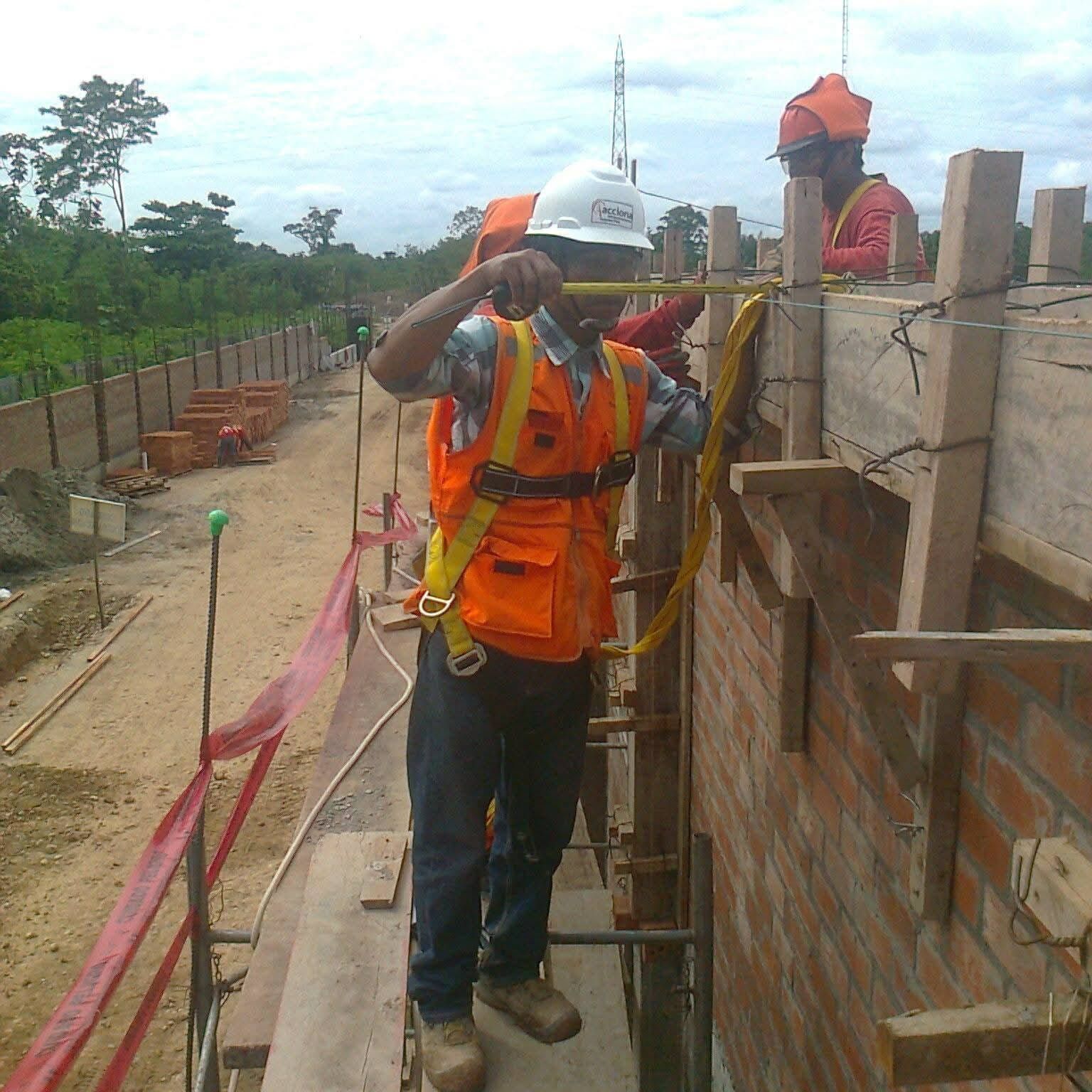 Un trabajador de la construcción con chaleco de seguridad naranja mide una pared de ladrillos en un andamio.