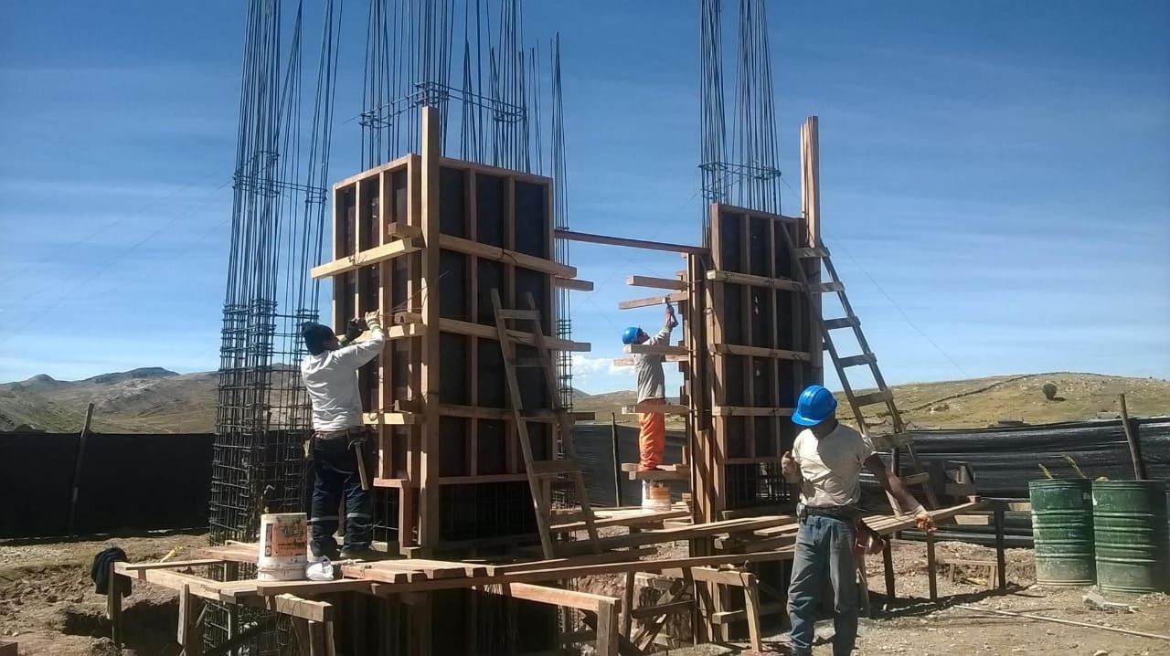 Obreros de la construcción construyen encofrados de hormigón al aire libre. Cielo azul y montañas al fondo.