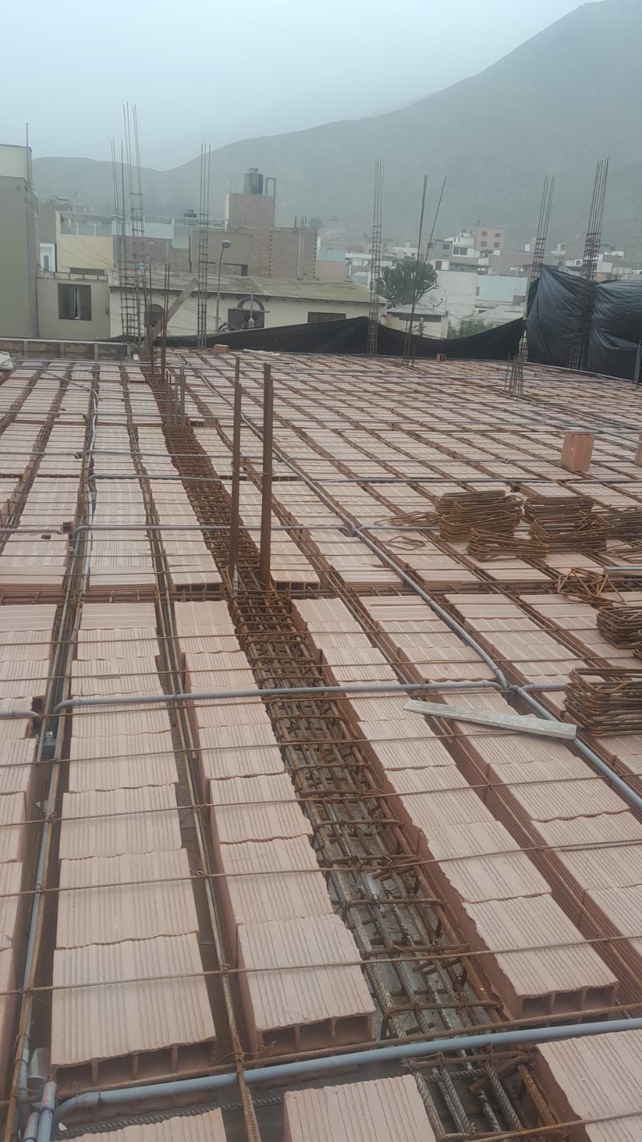 Obra: Rejilla de varillas corrugadas colocada sobre bloques de terracota en un tejado. Edificios y montañas al fondo.