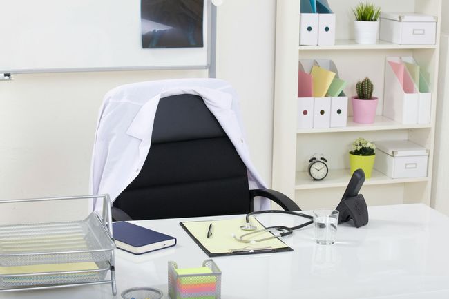 Bright office desk with a black chair, laptop, notebook, and shelves of colorful folders in the background