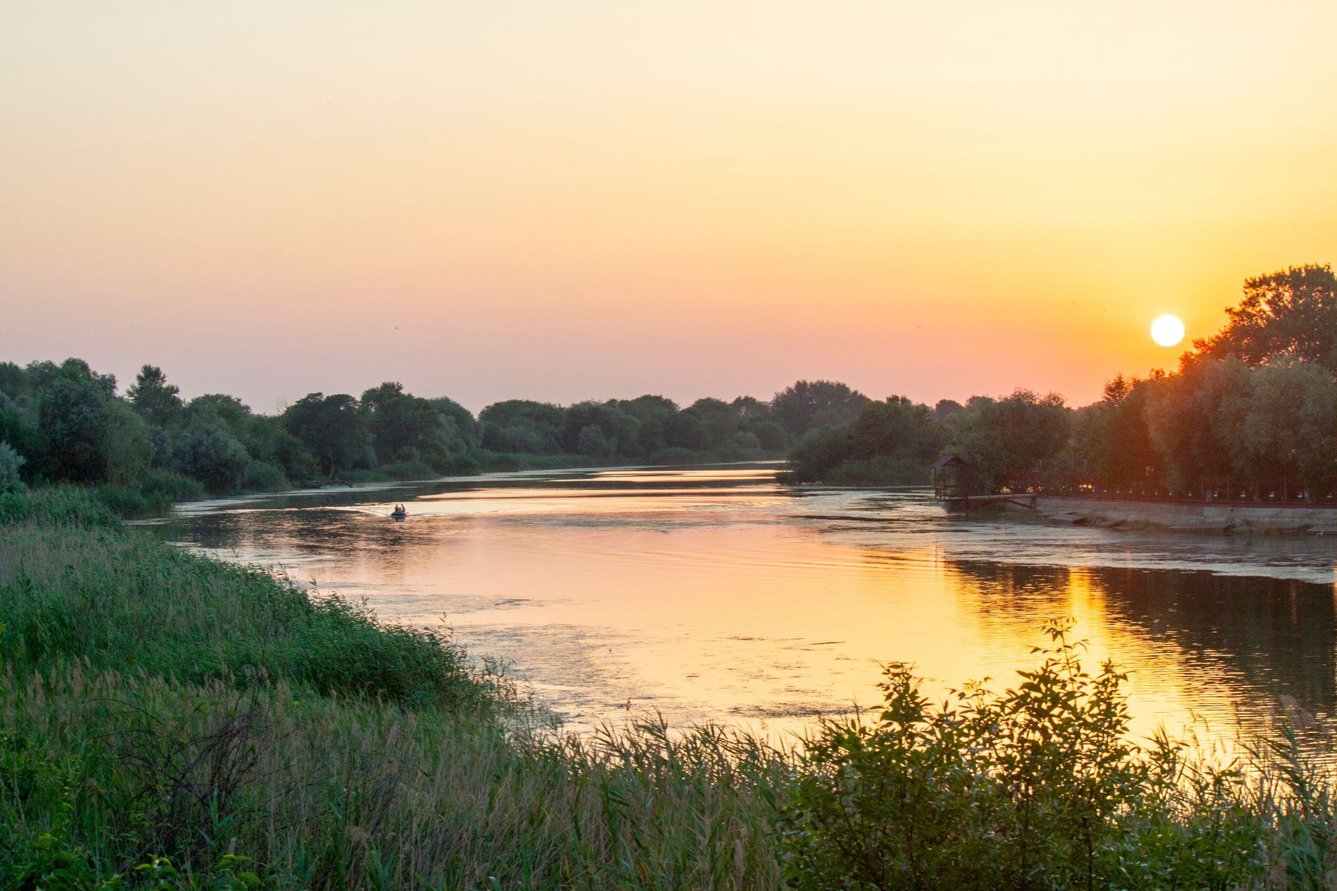 Sunset over a calm river, reflecting the orange and pink sky. Lush green trees line the banks.