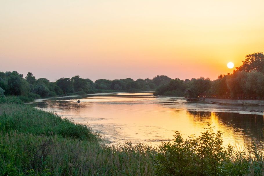 Sunset over a calm river reflecting the orange sky, with trees and green banks.