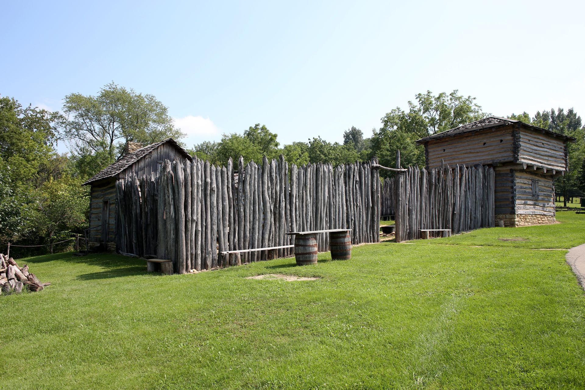 Wooden fort with a tall stockade fence, grass, and trees in the background under a blue sky.