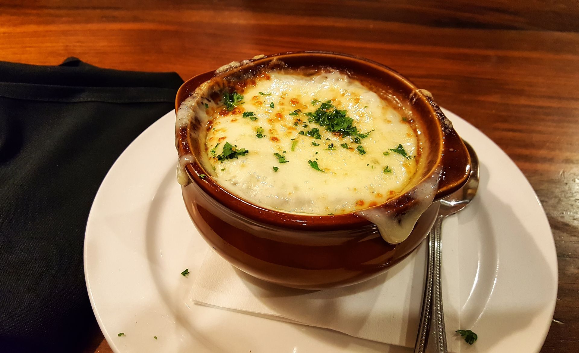 French onion soup in a brown bowl with melted cheese, parsley garnish, on a white plate.