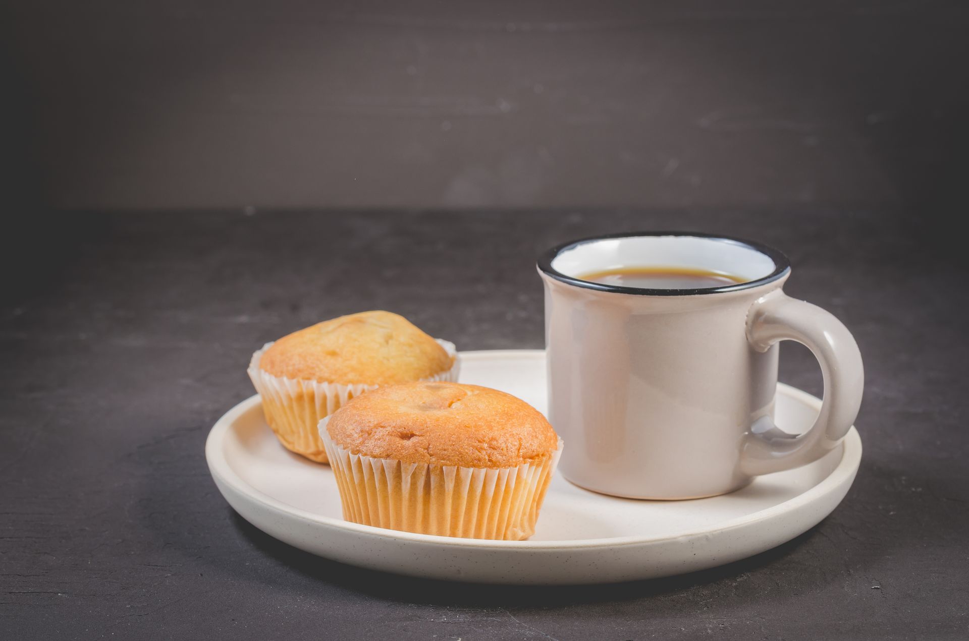 Two muffins and a cup of coffee on a white plate, set against a dark background.