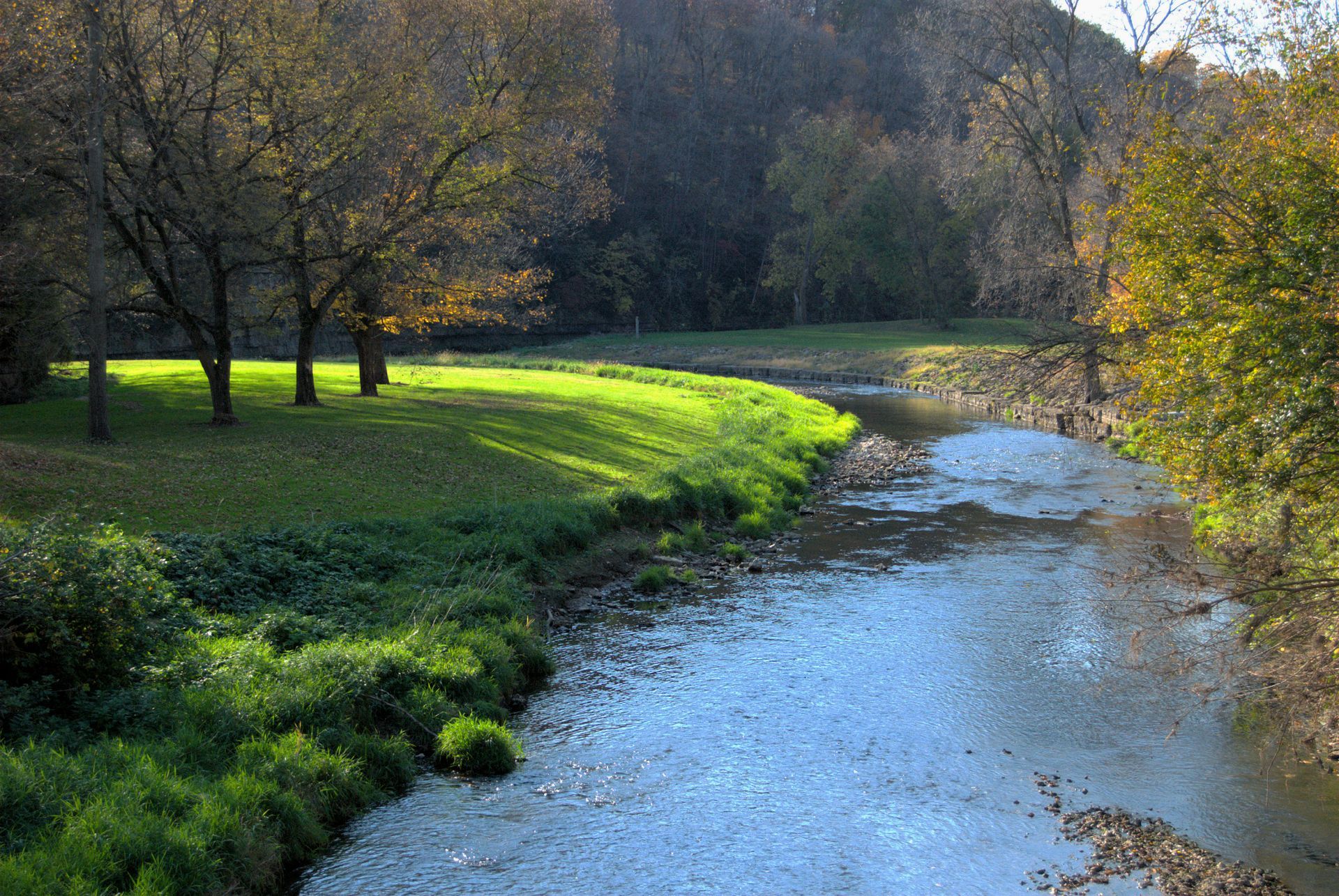 A sunny stream winds through a grassy bank and trees in autumn colors.
