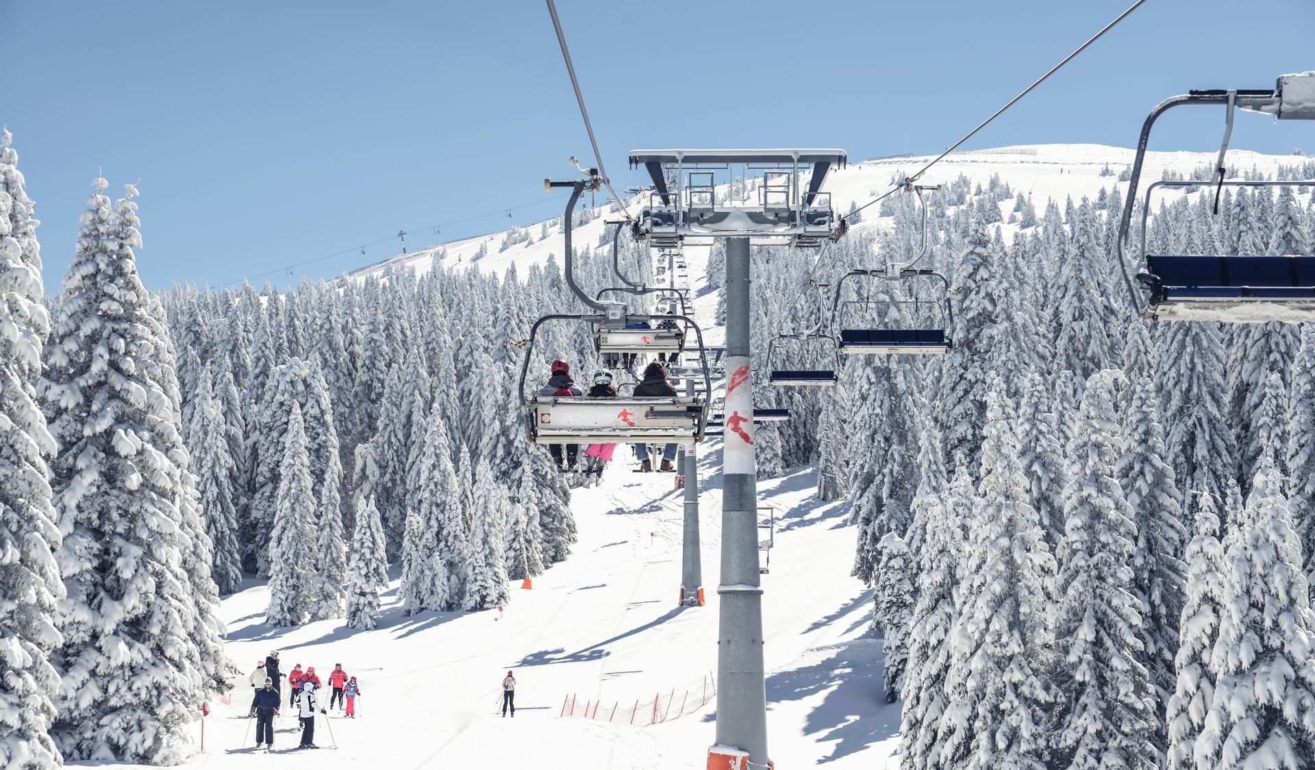 Ski lift carrying people up a snowy mountain slope, surrounded by snow-covered trees, under a bright blue sky.