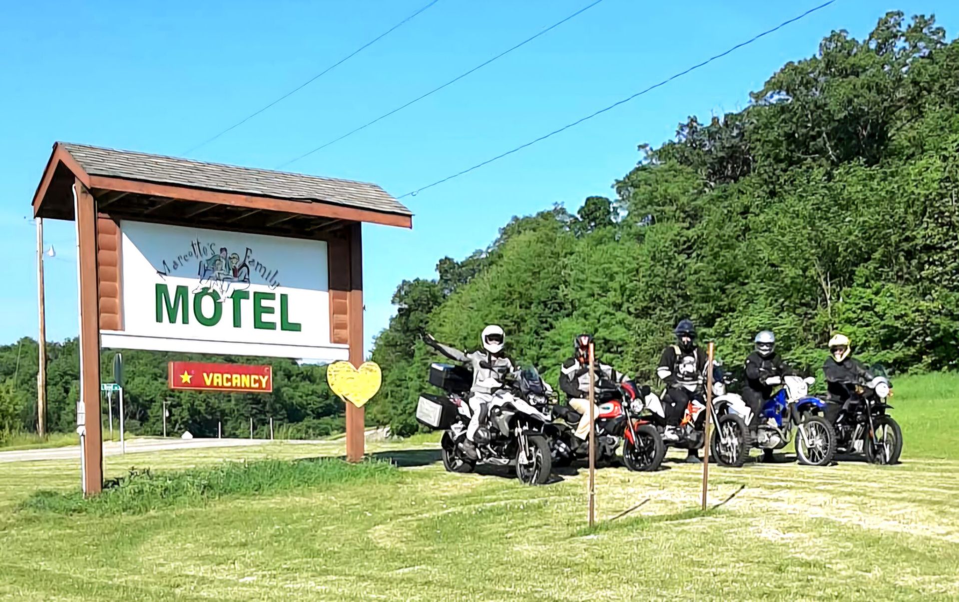 Motorcycles parked near a motel sign; riders in gear. Sunny day, green field and trees.