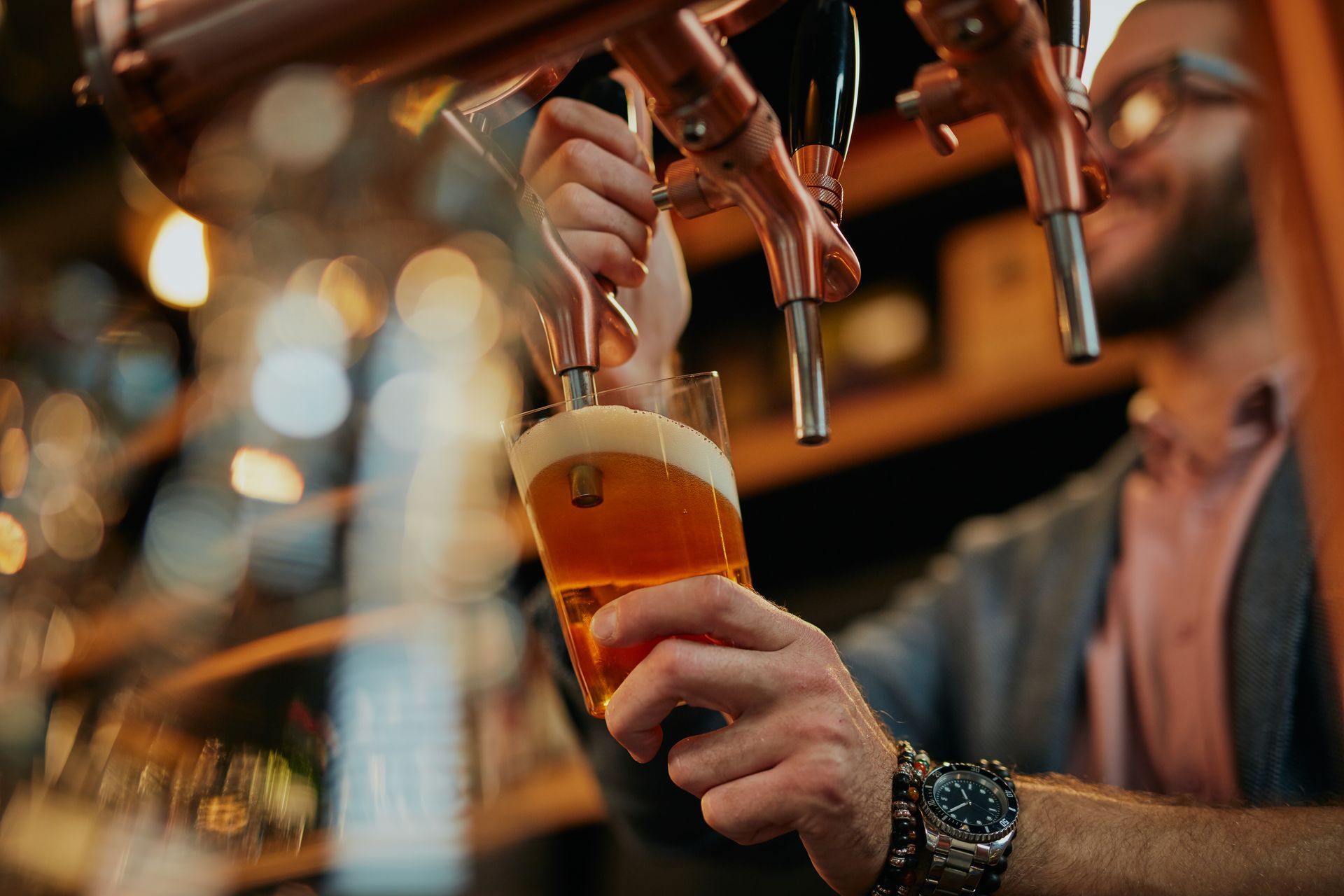 Hand pouring a beer into a glass at a bar, with warm blurred lights in the background