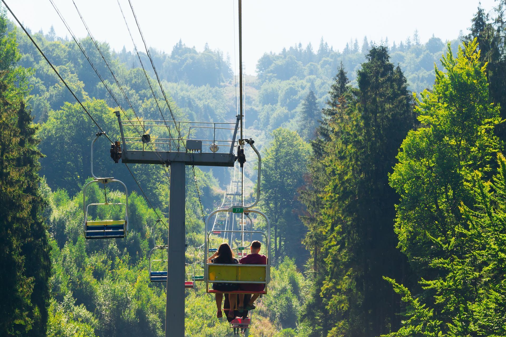 Chairlift with two riders gliding over a green forest on a sunny day