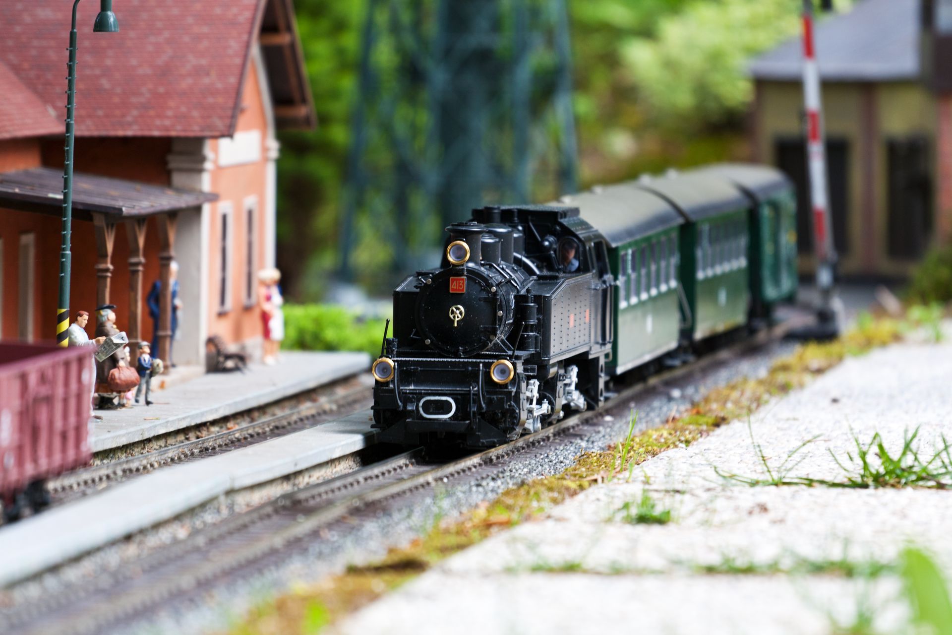A miniature steam train model traveling on tracks past a small station building with figures on the platform.