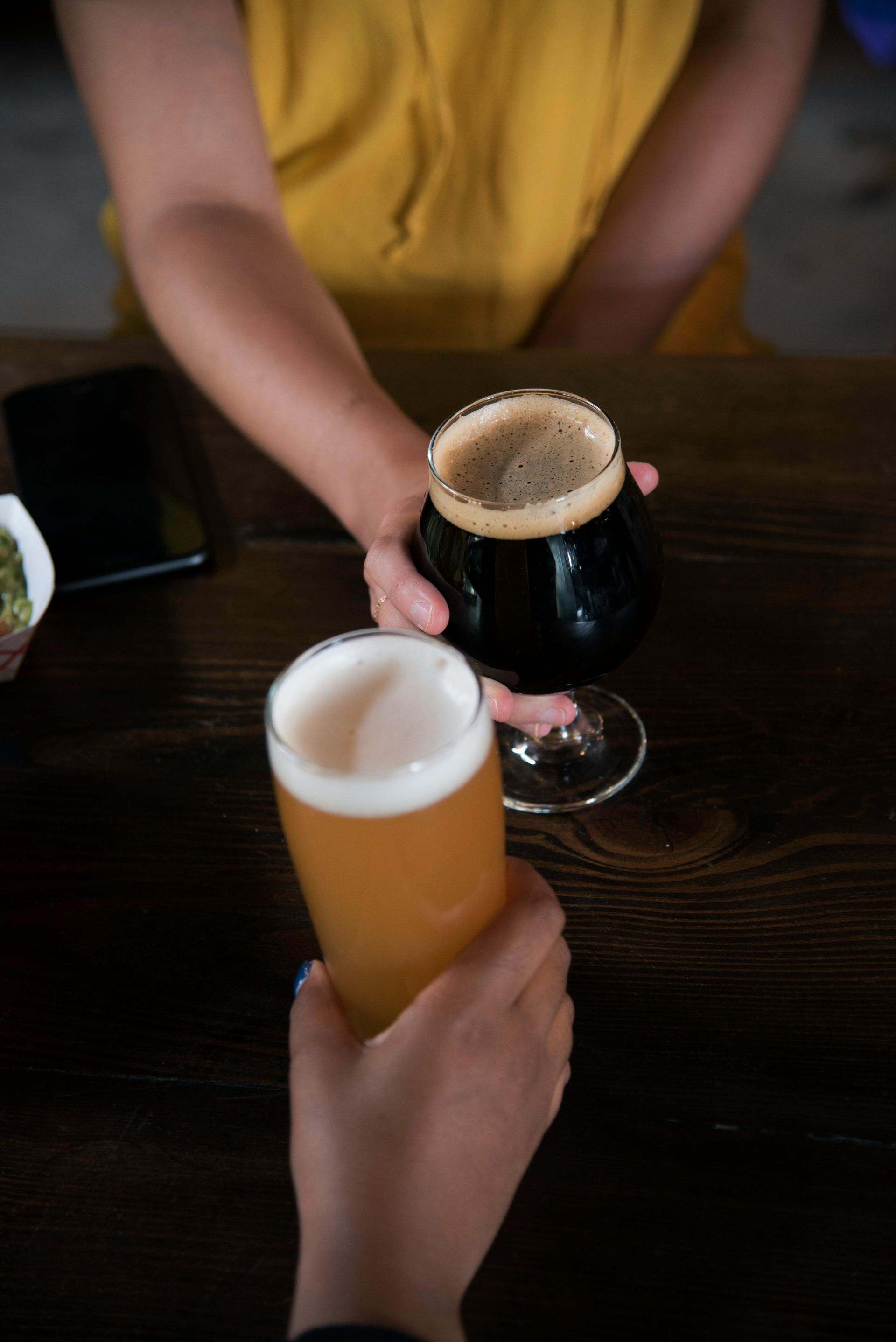 Two people toasting beer glasses at a dark wooden table. One holds dark beer, the other a light ale.