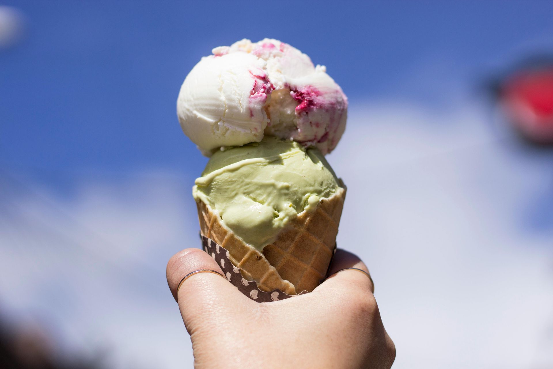 Hand holding a two-scoop ice cream cone: pistachio and berry flavors against a blue sky.