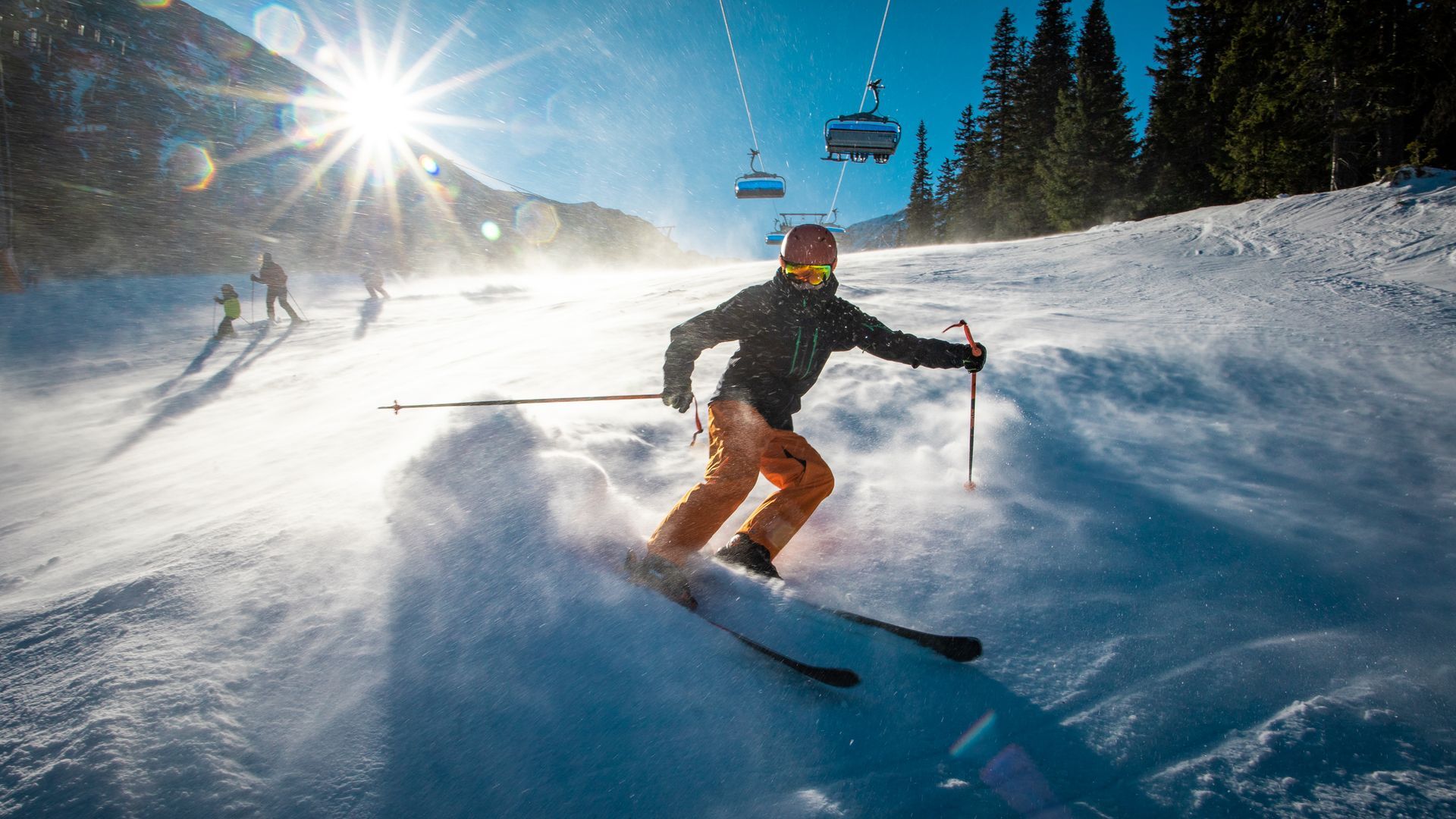 Skier carving through powder on a sunny mountain slope, with chairlifts and pine trees in the background