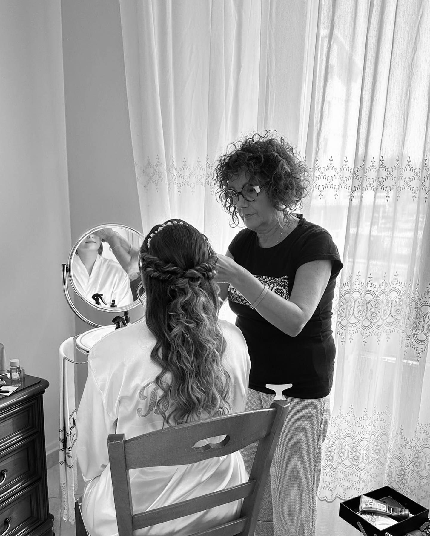 Donna che si fa acconciare i capelli da un'altra donna; vicino a una finestra e a uno specchio.