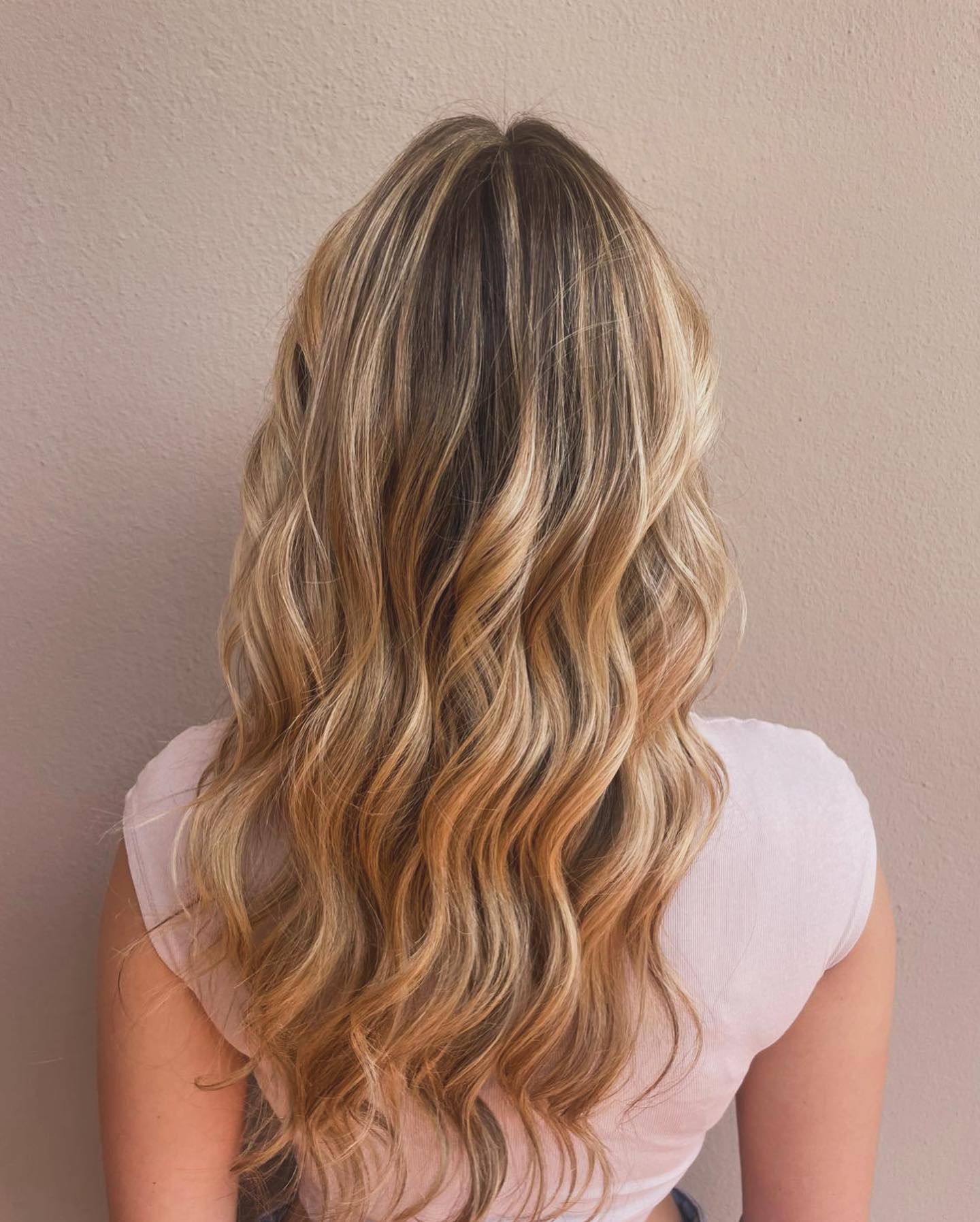 Donna con capelli biondi lunghi e ondulati, mèches e radici scure. Top rosa chiaro su sfondo neutro.