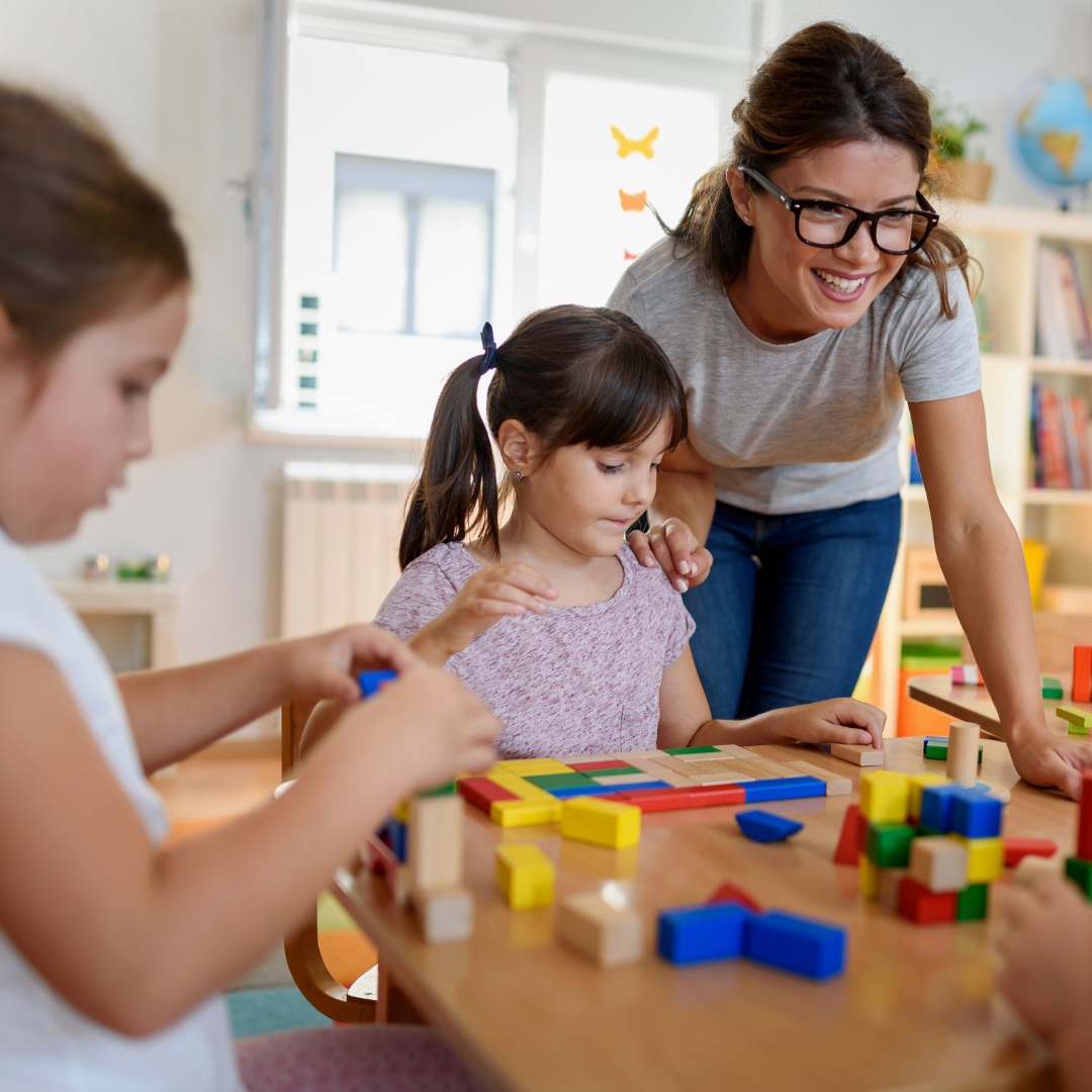Teacher assisting two young girls with building blocks at a table in a brightly lit classroom.