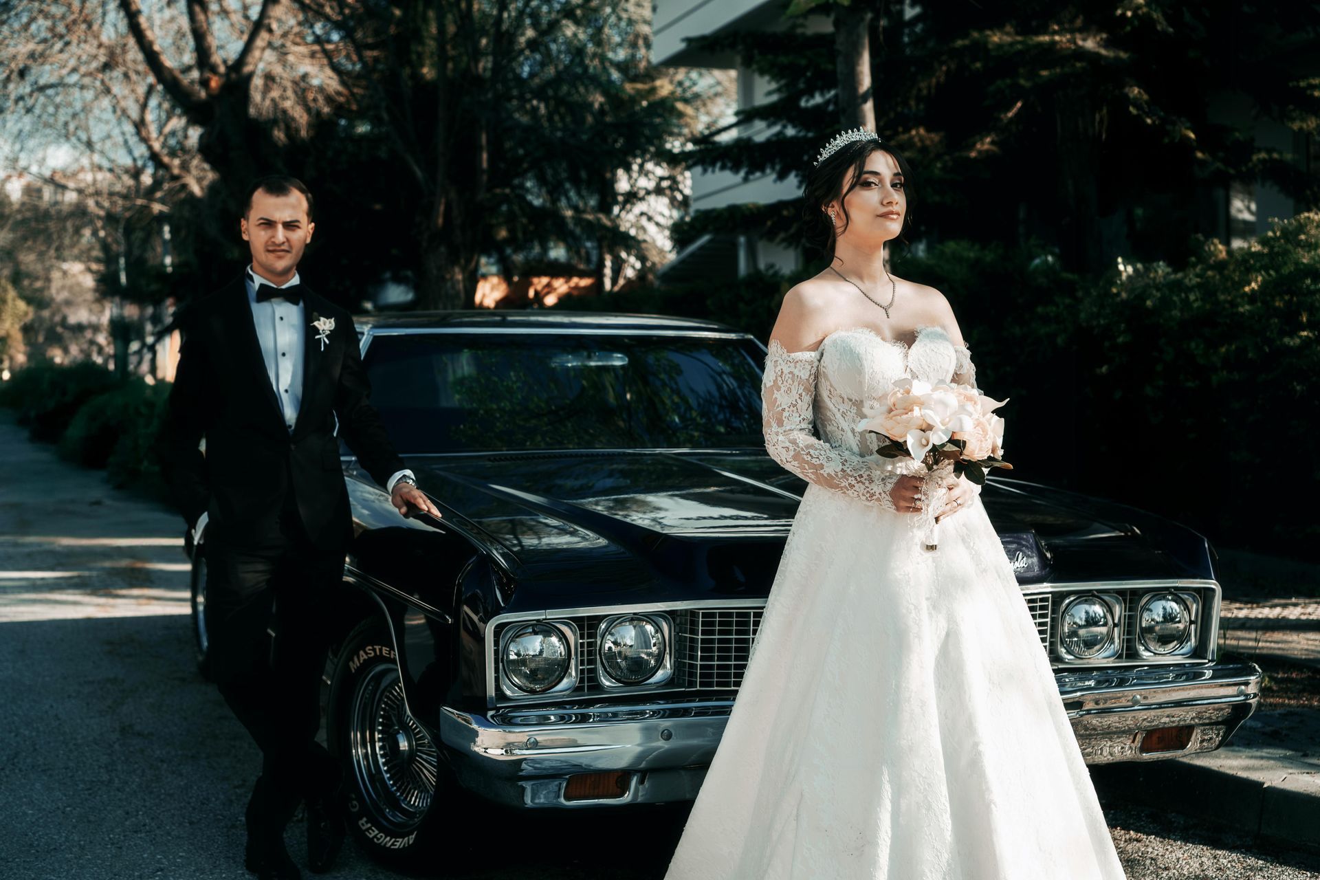 Bride and groom posing by a vintage black car on a tree-lined street; the bride holds a bouquet.
