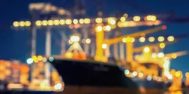 Blurred nighttime view of a cargo ship docked at a port, lit by bright yellow lights.