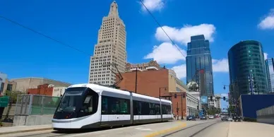A modern streetcar travels down a city street with skyscrapers in the background under a blue sky.