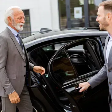 An older man in a suit is being assisted into a black car by another man, outdoors.
