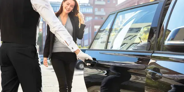 A chauffeur opening a car door for a smiling woman in front of a city building.