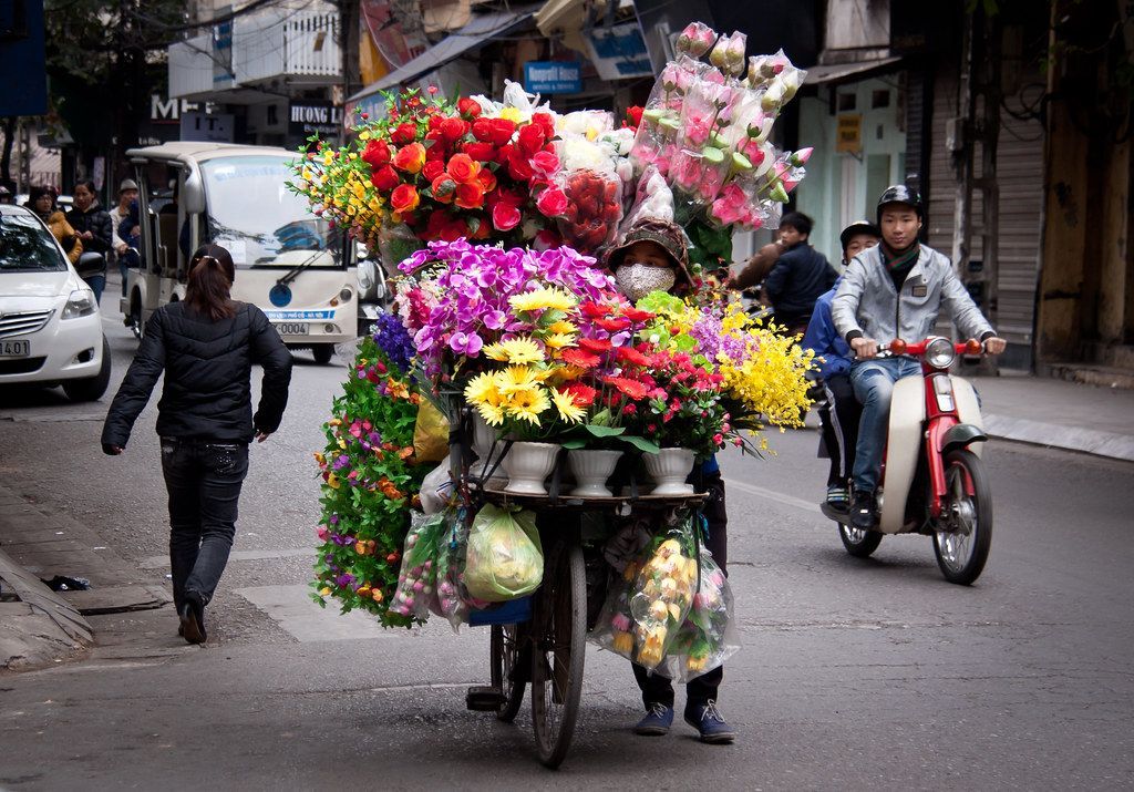 A street vendor rides a bicycle loaded with bright, multicolored flowers, almost covering their entire body, while navigating a busy city street.