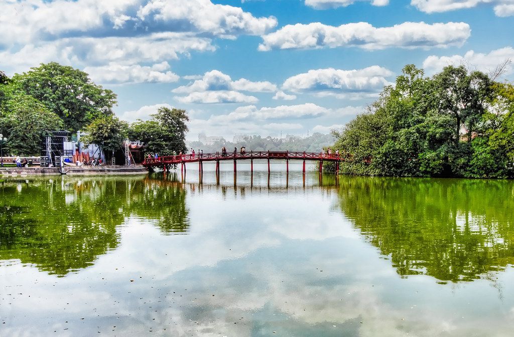A red wooden bridge stretches across a calm green lake surrounded by trees, with people walking across it under a bright blue sky with scattered clouds.