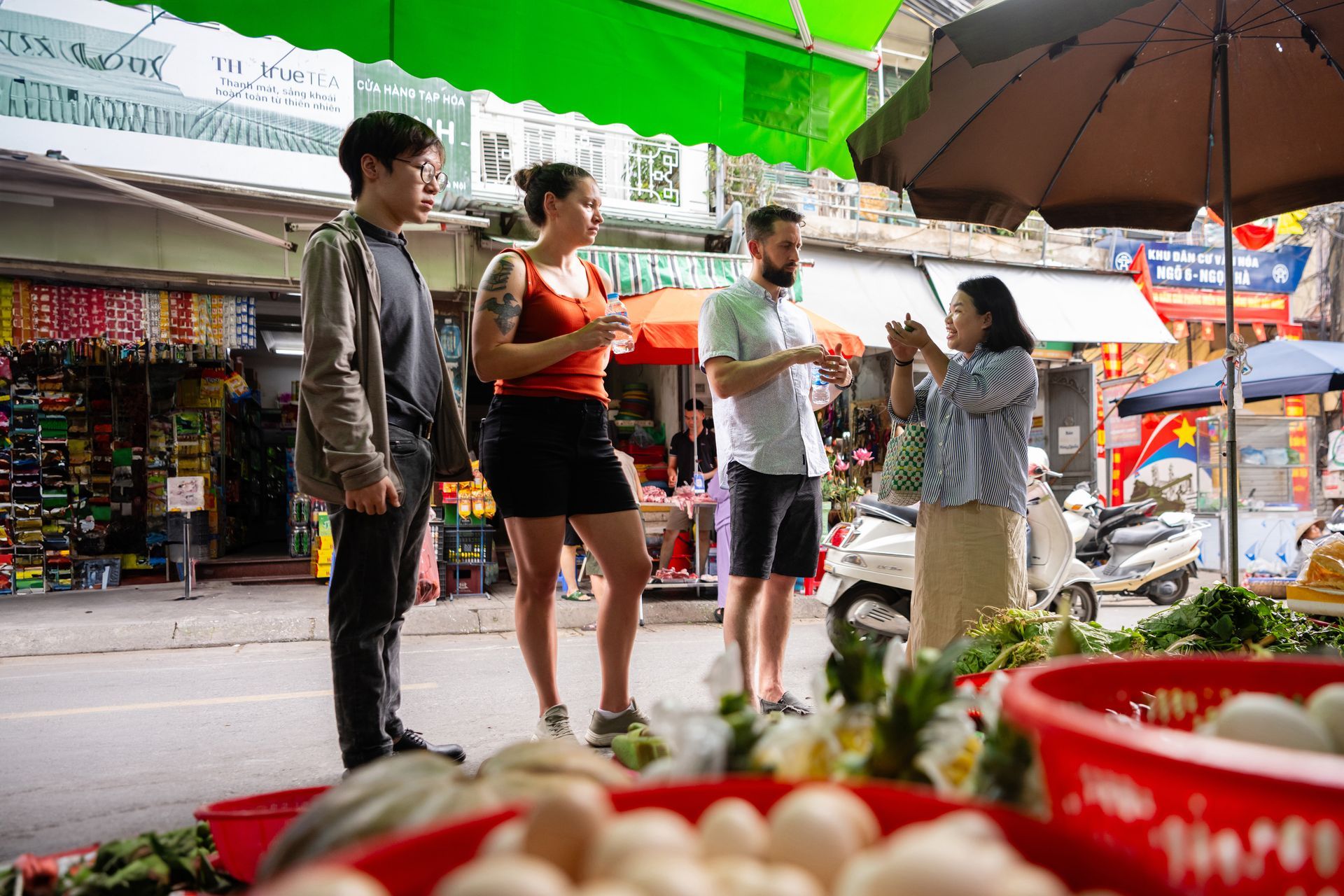 Two Vietnamese women wearing traditional conical hats exchanging goods from their bicycles loaded with fresh produce on a quiet street.