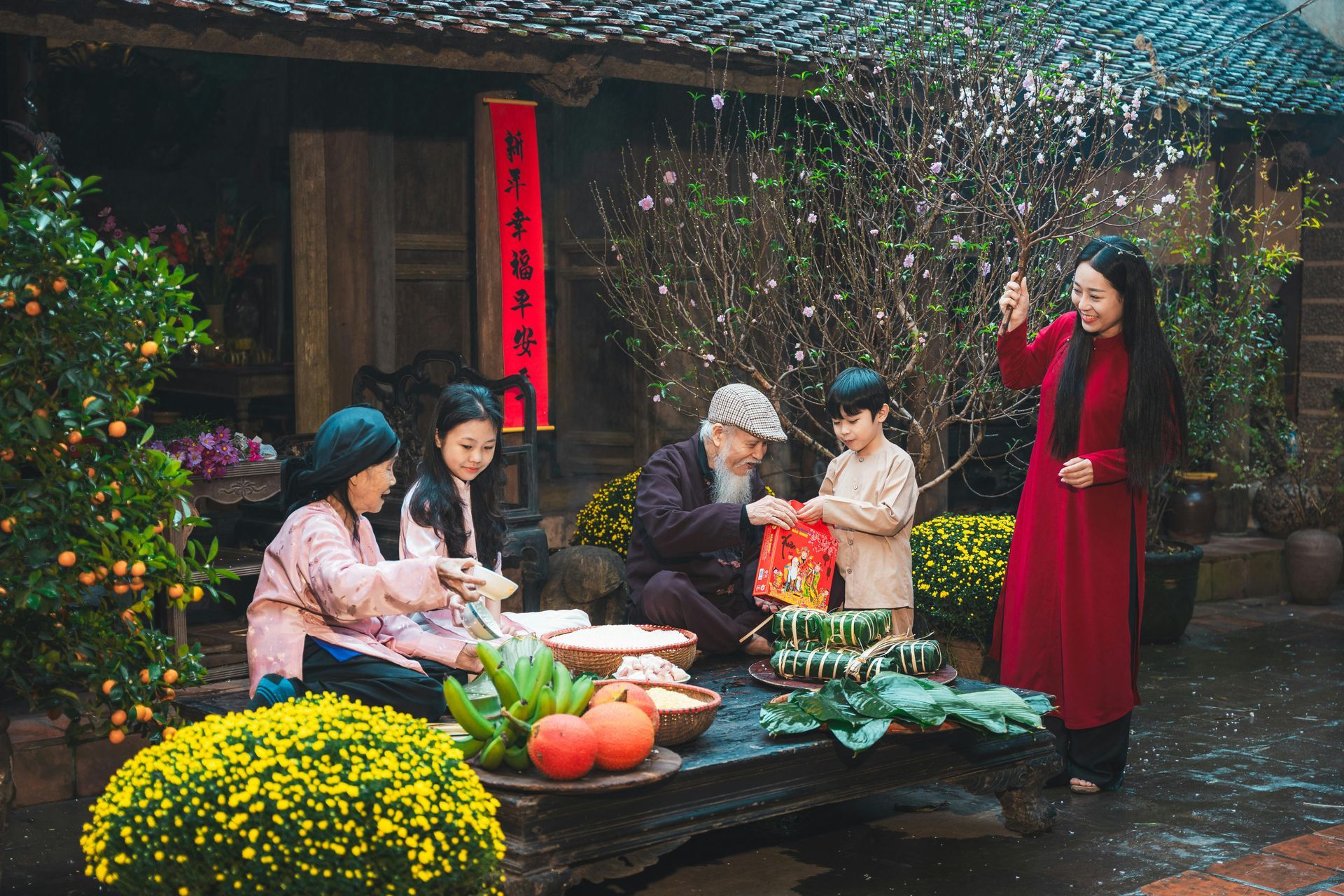 A Vietnamese family of multiple generations gathers in front of a traditional wooden house during Tết, surrounded by peach blossoms, kumquat trees, and festive food like bánh chưng and fruits. An elderly man gives a red envelope to a child while others prepare offerings.