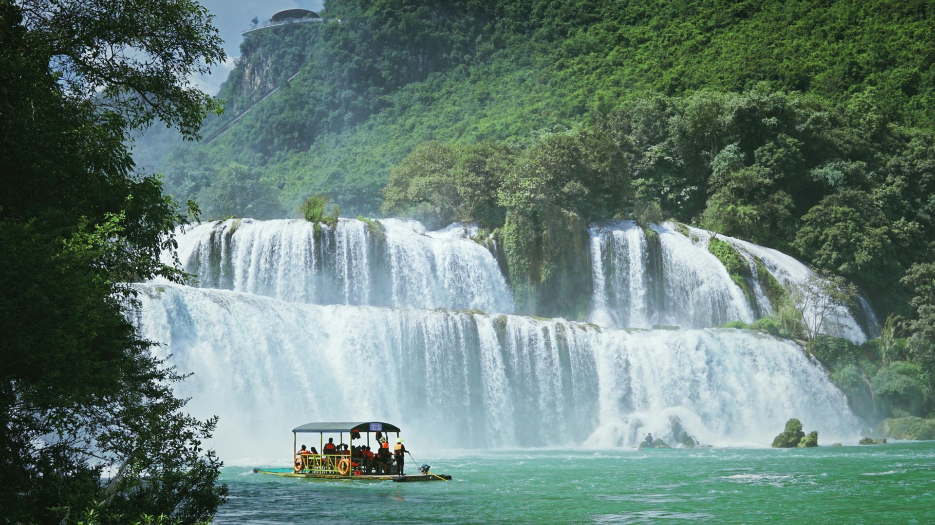 A serene scene of a boat floating near the Ban Gioc Waterfall, surrounded by green hills.