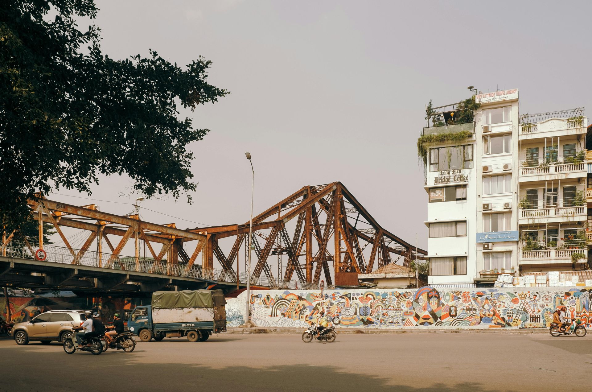 The historic Long Bien Bridge with its iron structure, alongside a colorful mural wall in Hanoi; motorbikes and cars pass below.