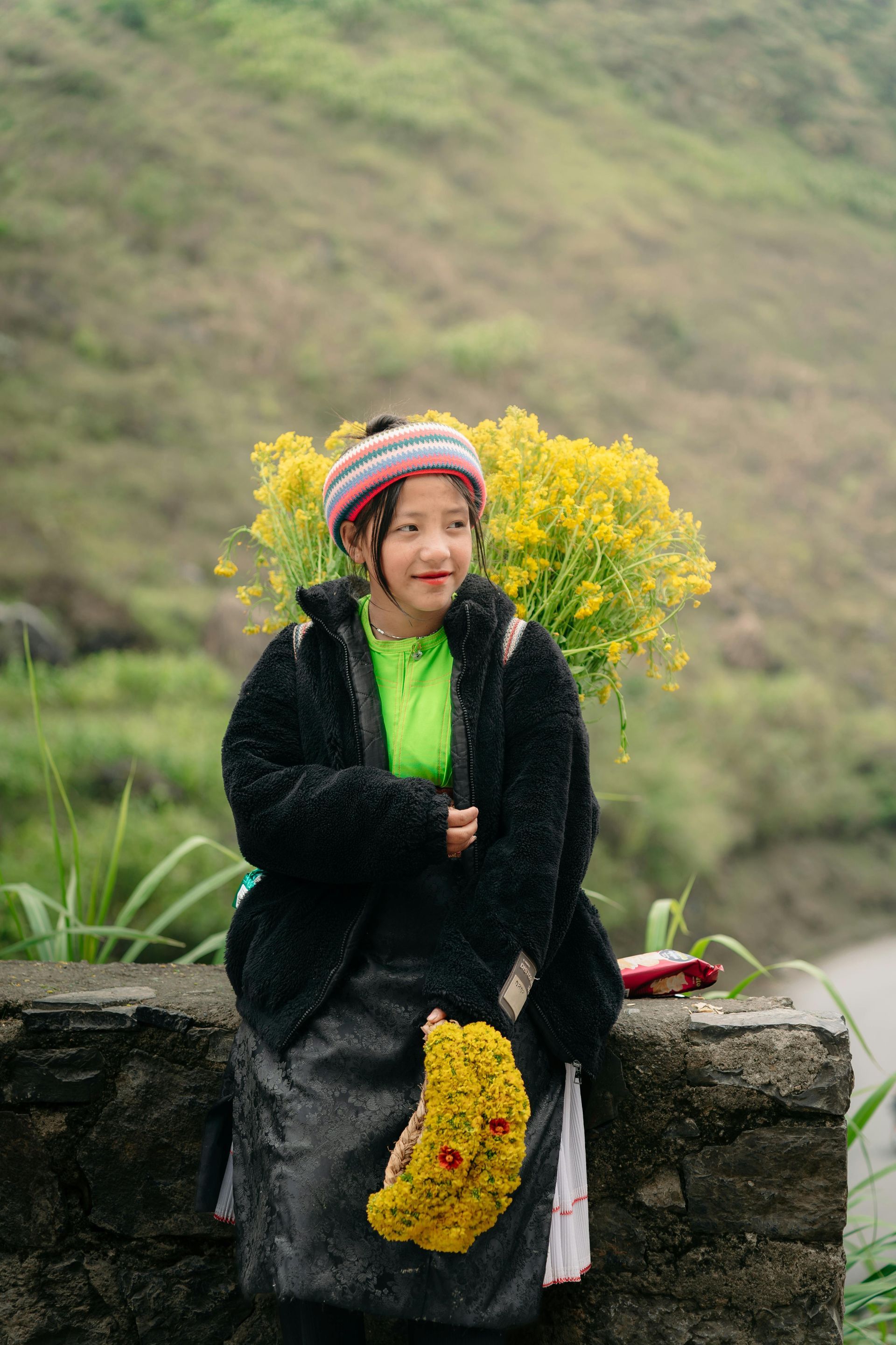 A little ethnic minority girl in Ha Giang carrying a basket of flowers on her back, holding handmade floral wreaths in her hands.