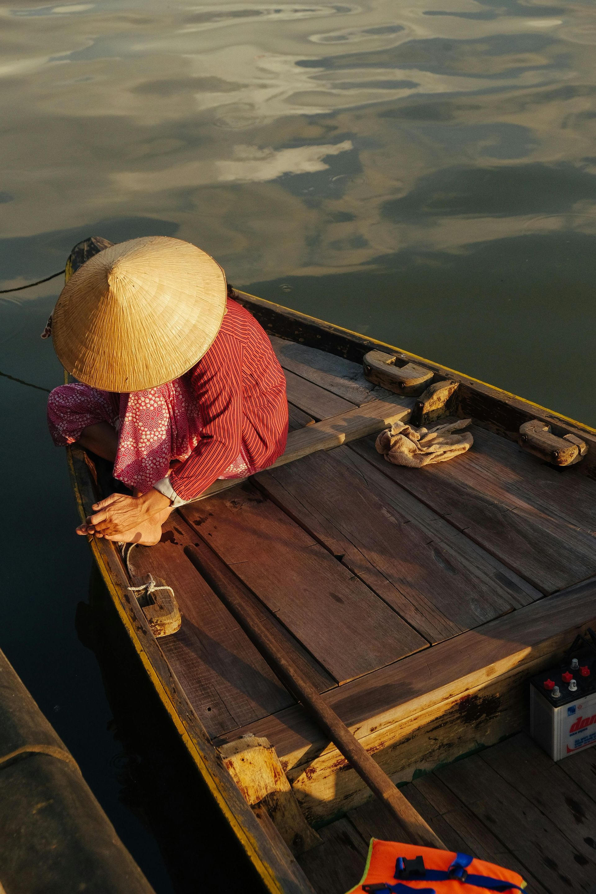 A woman wearing a traditional Vietnamese conical hat (nón lá) sitting on a wooden boat, gently floating on a calm river.