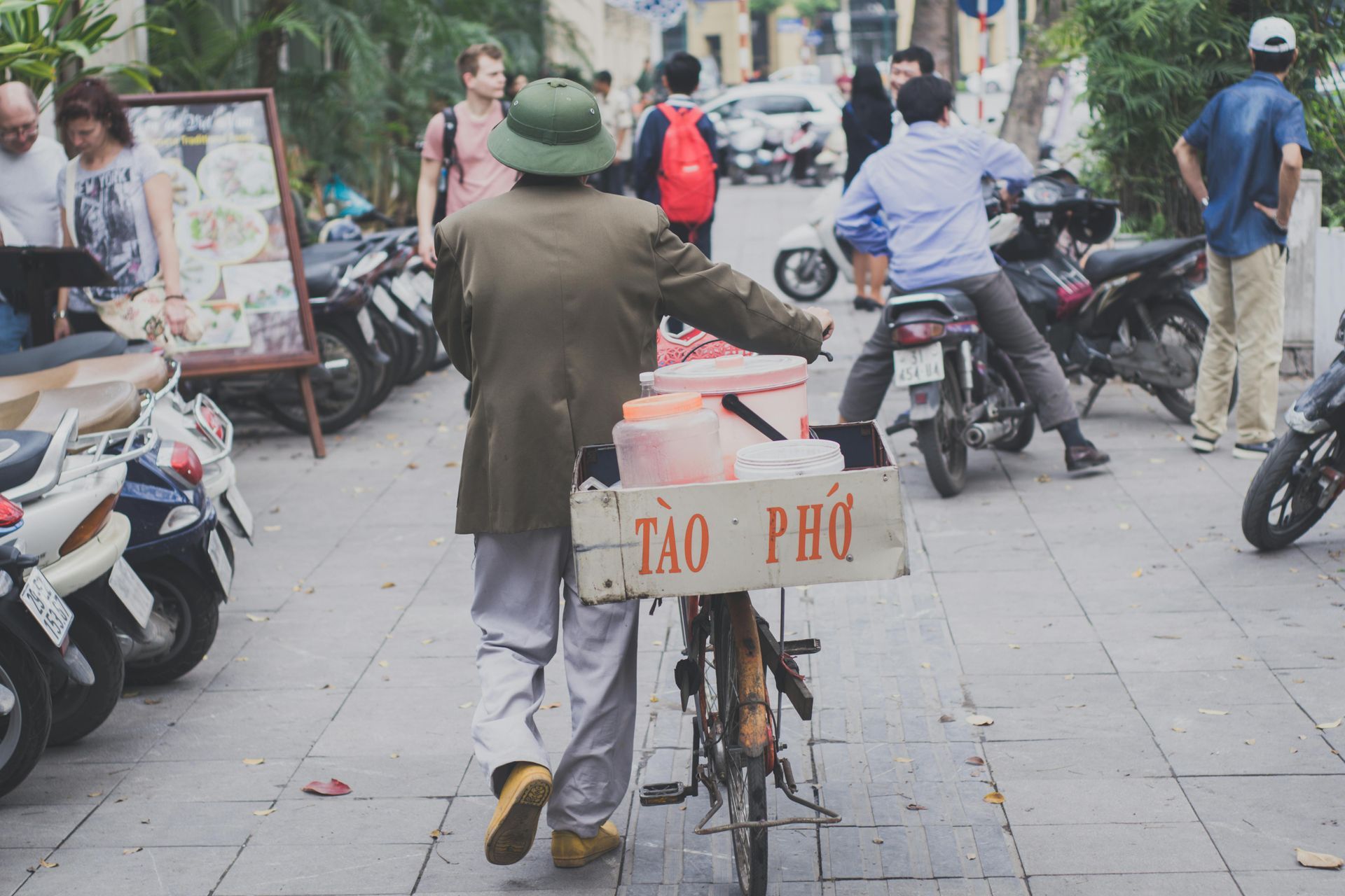 A man wearing an army green jacket and a pith helmet, pushing a bicycle carrying a box labeled 