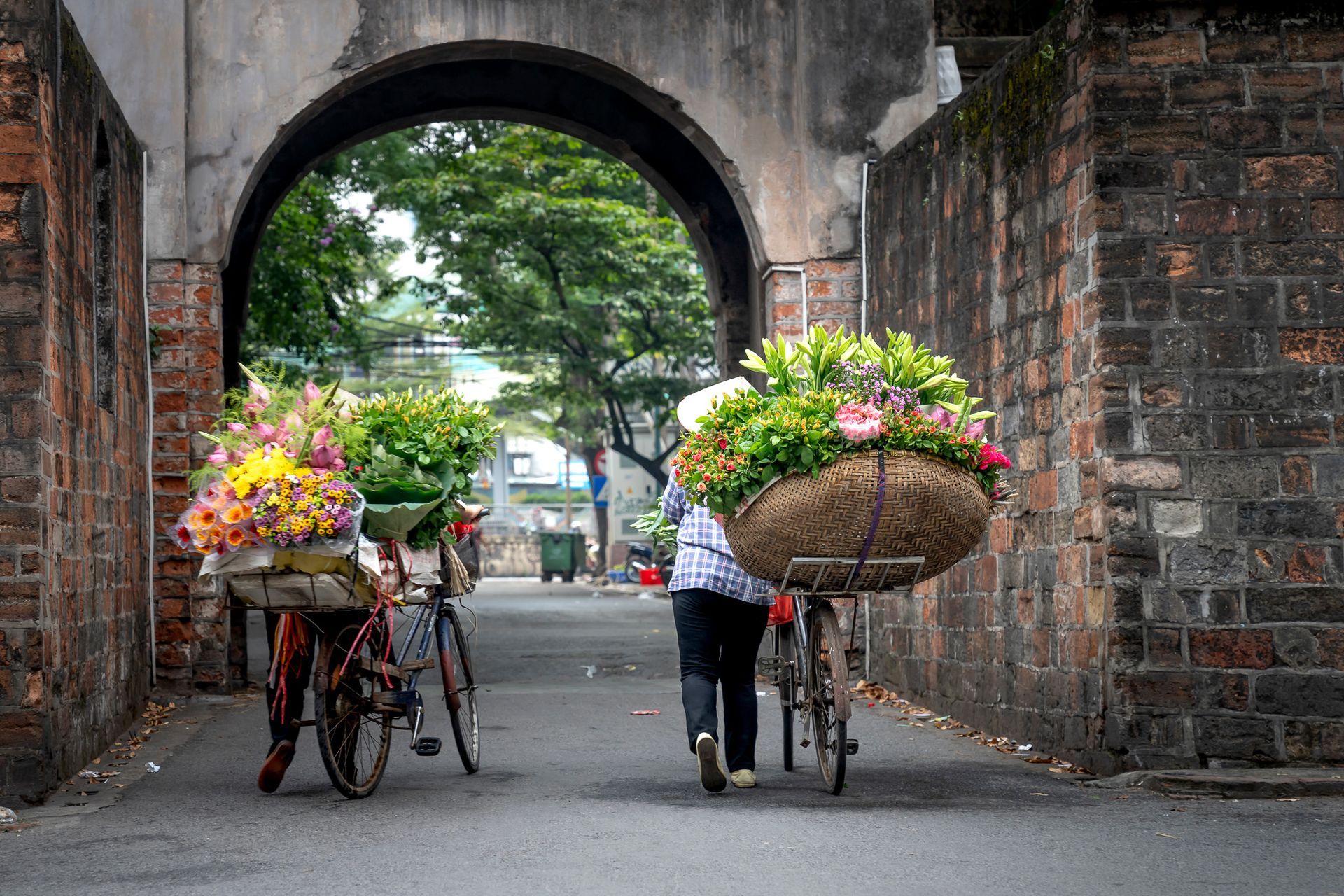 Two vendors pushing bicycles loaded with colorful flowers through an ancient stone gate in Vietnam.