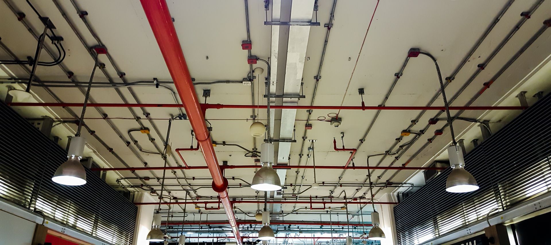 Industrial ceiling with exposed red pipes, ducts, and hanging lights in a long corridor.
